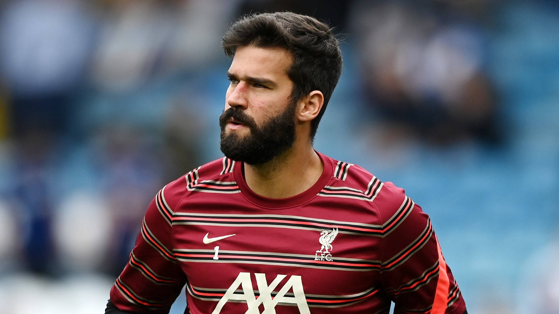 Alisson Becker 桌布 壁紙 — 一位專注的足球守門員穿著紅色條紋球衣，站在球場上，身後是模糊的藍色座位。