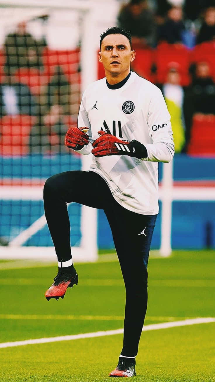 Keylor Navas 桌布 壁紙 — 一位專注的足球守門員身穿白色訓練服，在充滿活力的綠色球場上伸展，背景中的體育場座位模糊不清。
