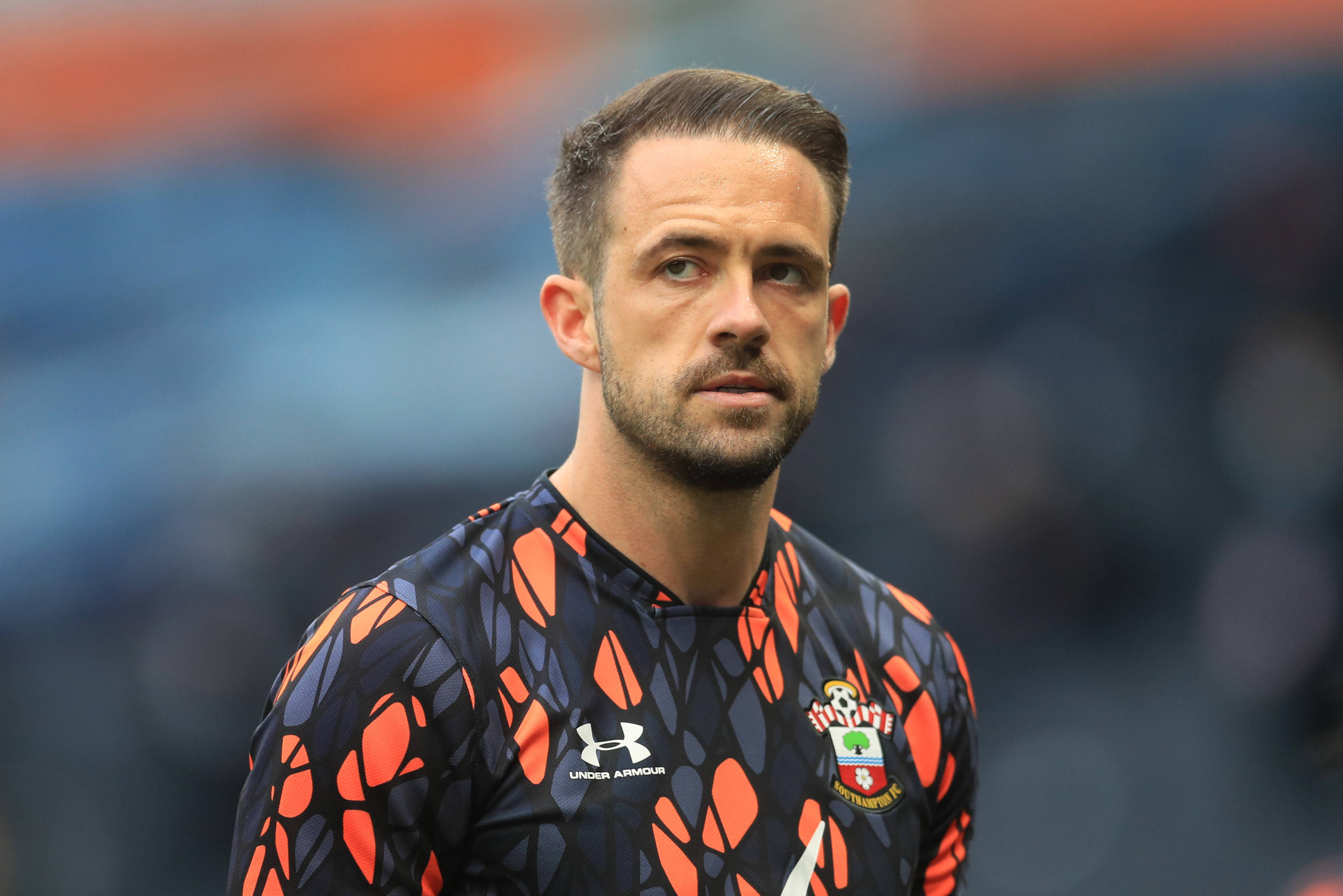 Football Players 桌布 壁紙 — 一位專注的足球守門員身穿黑色和橙色圖案球衣，站在模糊的球場上。
