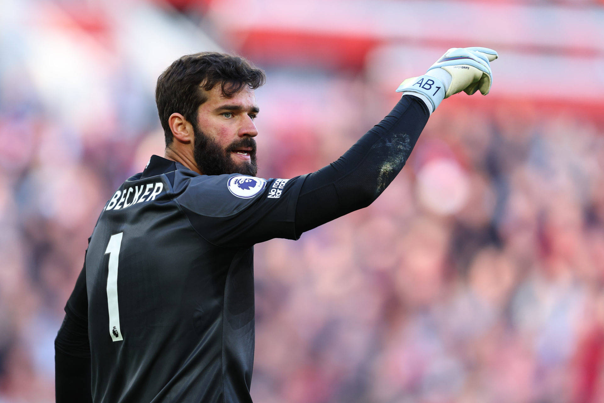 Alisson Becker 桌布 壁紙 — 一位專注的足球守門員身穿黑色球衣，背號1，在日間比賽中強烈地做手勢。