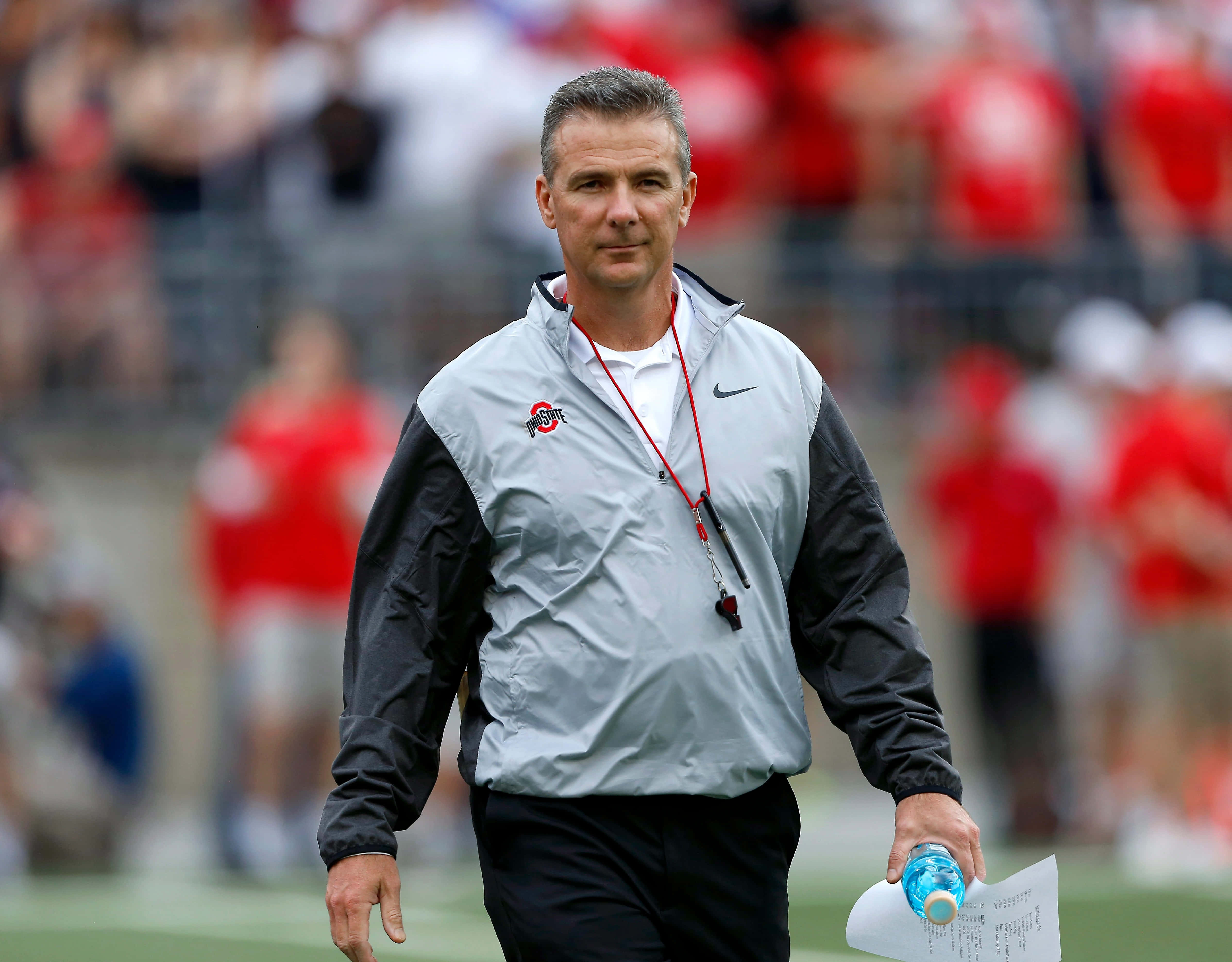 Urban Meyer 桌布 壁紙 — 一位專注的足球教練穿著灰色和黑色外套，拿著水壺和文件在綠色球場上行走。