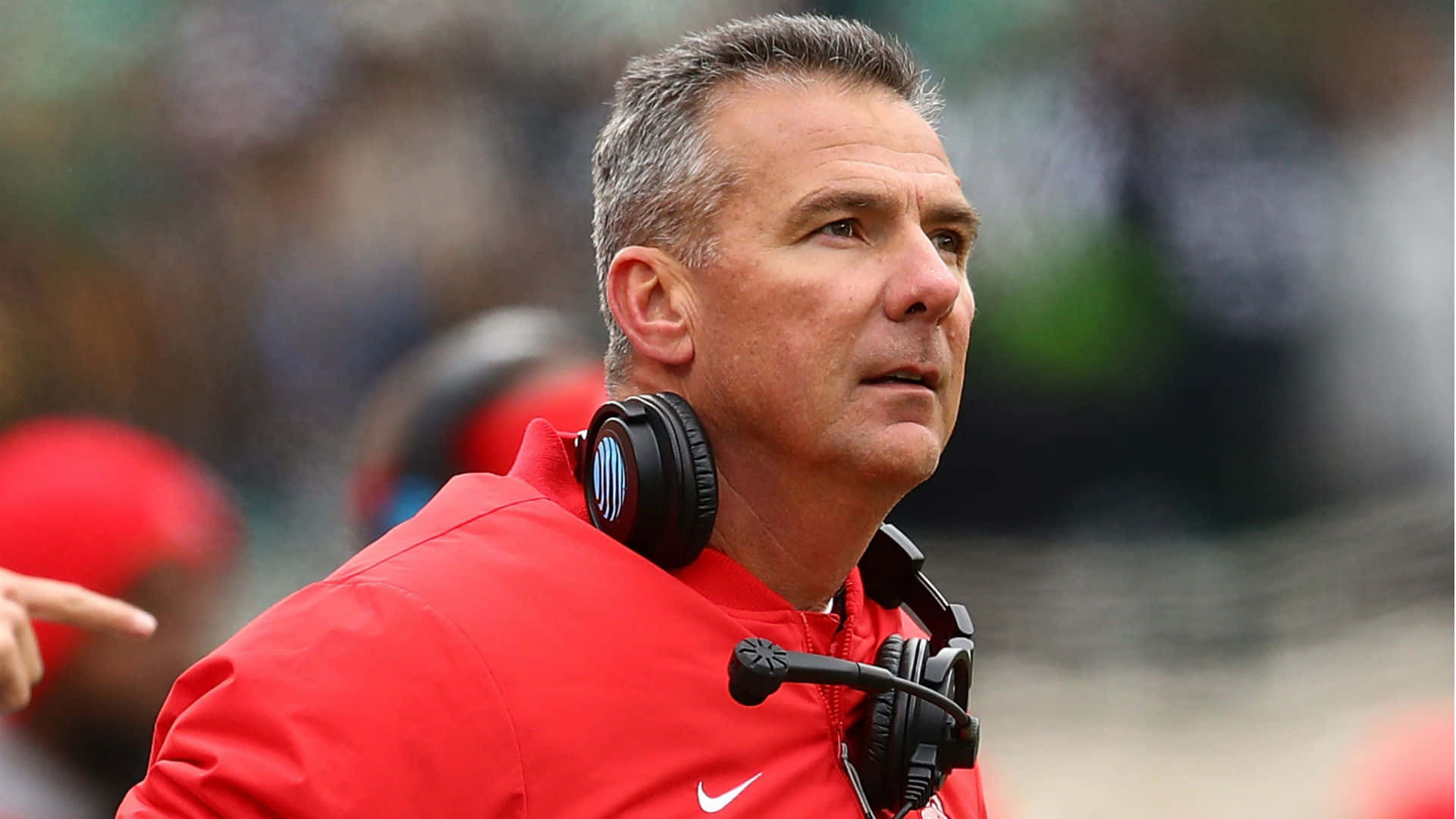 Urban Meyer 桌布 壁紙 — 一位專注的足球教練穿著紅色外套和耳機，在比賽中專注地看著球場。