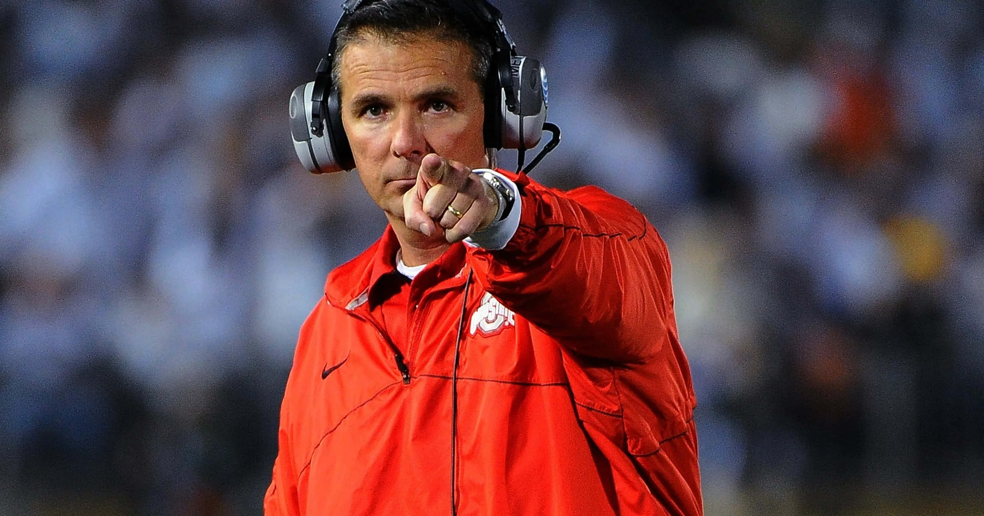 Urban Meyer 桌布 壁紙 — 一位專注的足球教練穿著紅色外套和耳機，在夜間比賽中在球場燈光下專注地指點。