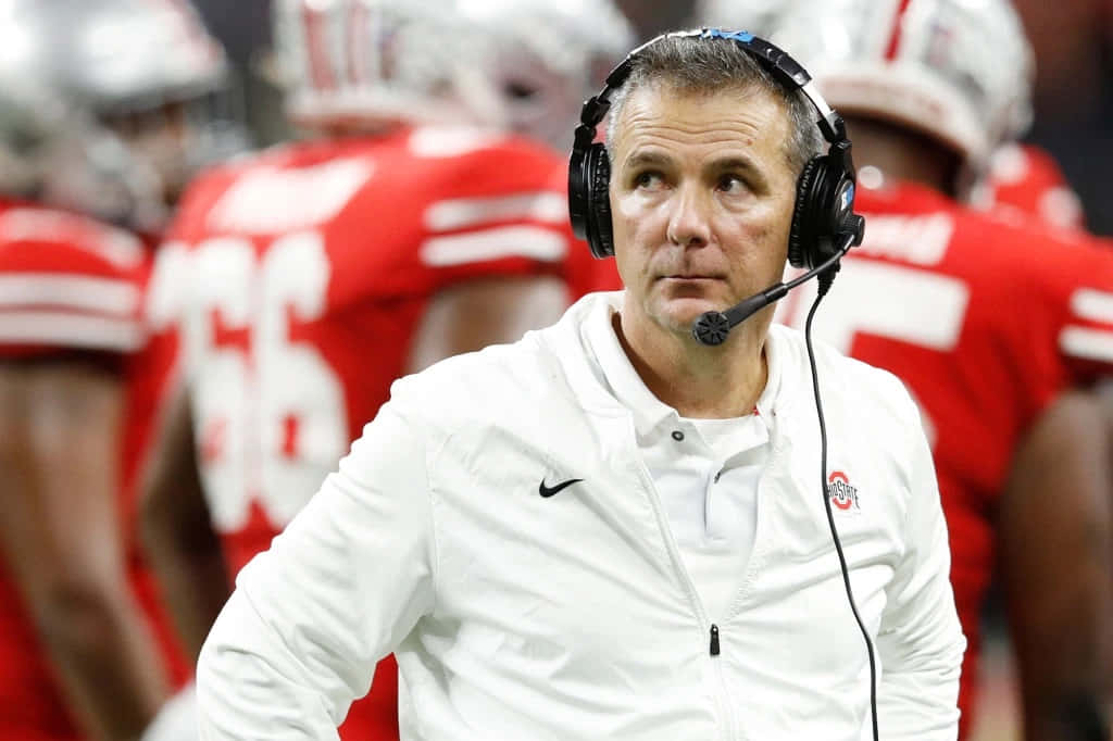 Urban Meyer 桌布 壁紙 — 一位專注的足球教練身穿白色外套和耳機，站在邊線，身後有穿著紅色球衣的球員模糊背景。