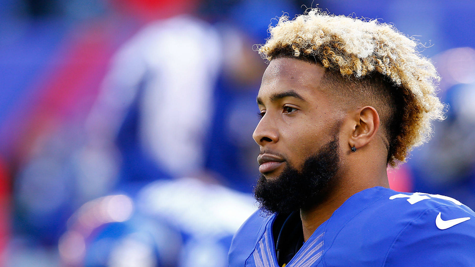 Odell 桌布 壁紙 — 一位專注的足球運動員，金色捲髮和鬍鬚，身穿藍色球衣，背景是模糊的體育場。