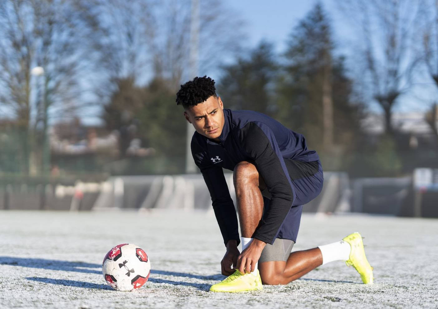 Ollie Watkins 桌布 壁紙 — 一位專注的足球運動員跪在結霜的草地上，在紅白球旁邊繫著亮黃色的球鞋，天空清澈湛藍。