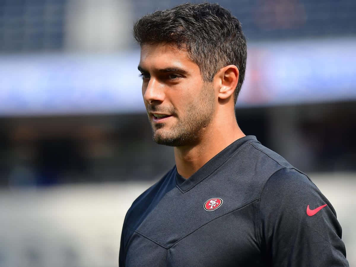 Jimmy Garoppolo 桌布 壁紙 — 一位專注的足球運動員身穿深色訓練球衣，帶有紅色標誌，站在陽光明媚的球場上，背景中的體育場座位模糊不清。