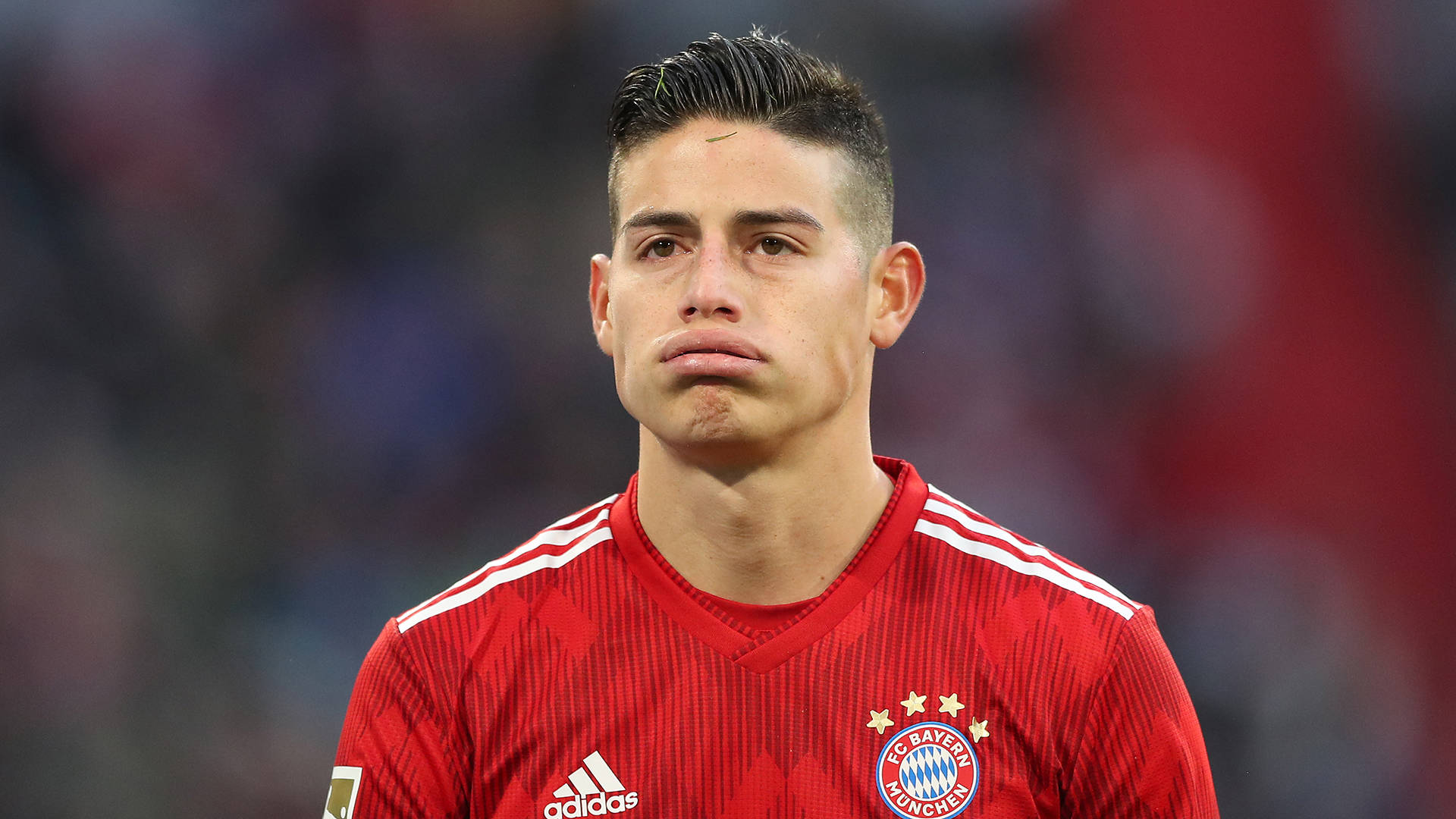 James Rodriguez 桌布 壁紙 — 一位專注的足球運動員身穿紅色拜仁慕尼黑球衣，在模糊的球場背景下帶著嚴肅的表情站立。