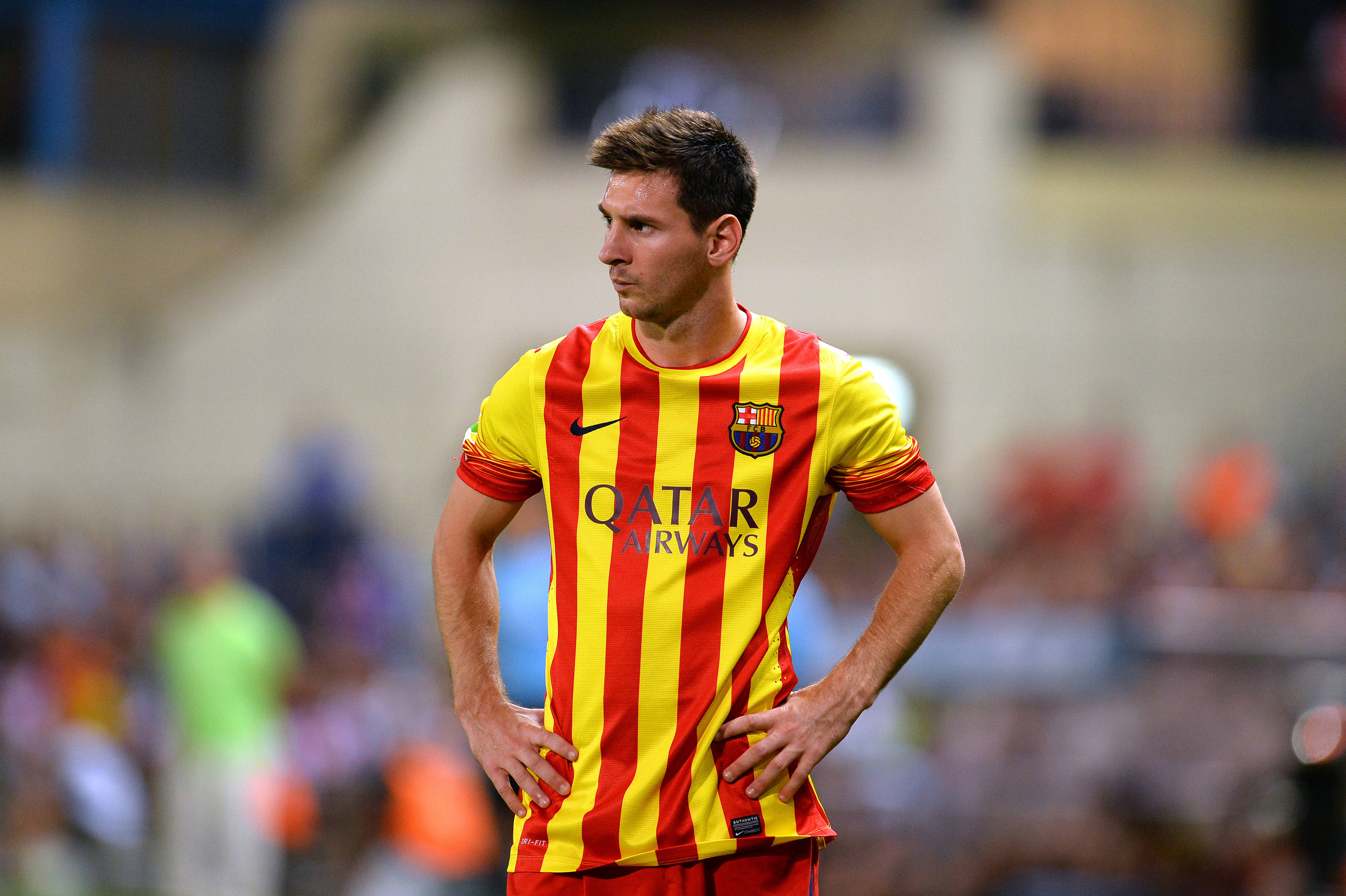 Messi 4K Ultra HD 桌布 壁紙 — 一位專注的足球運動員身穿紅黃條紋球衣，在明亮的燈光下雙手叉腰站在球場上。