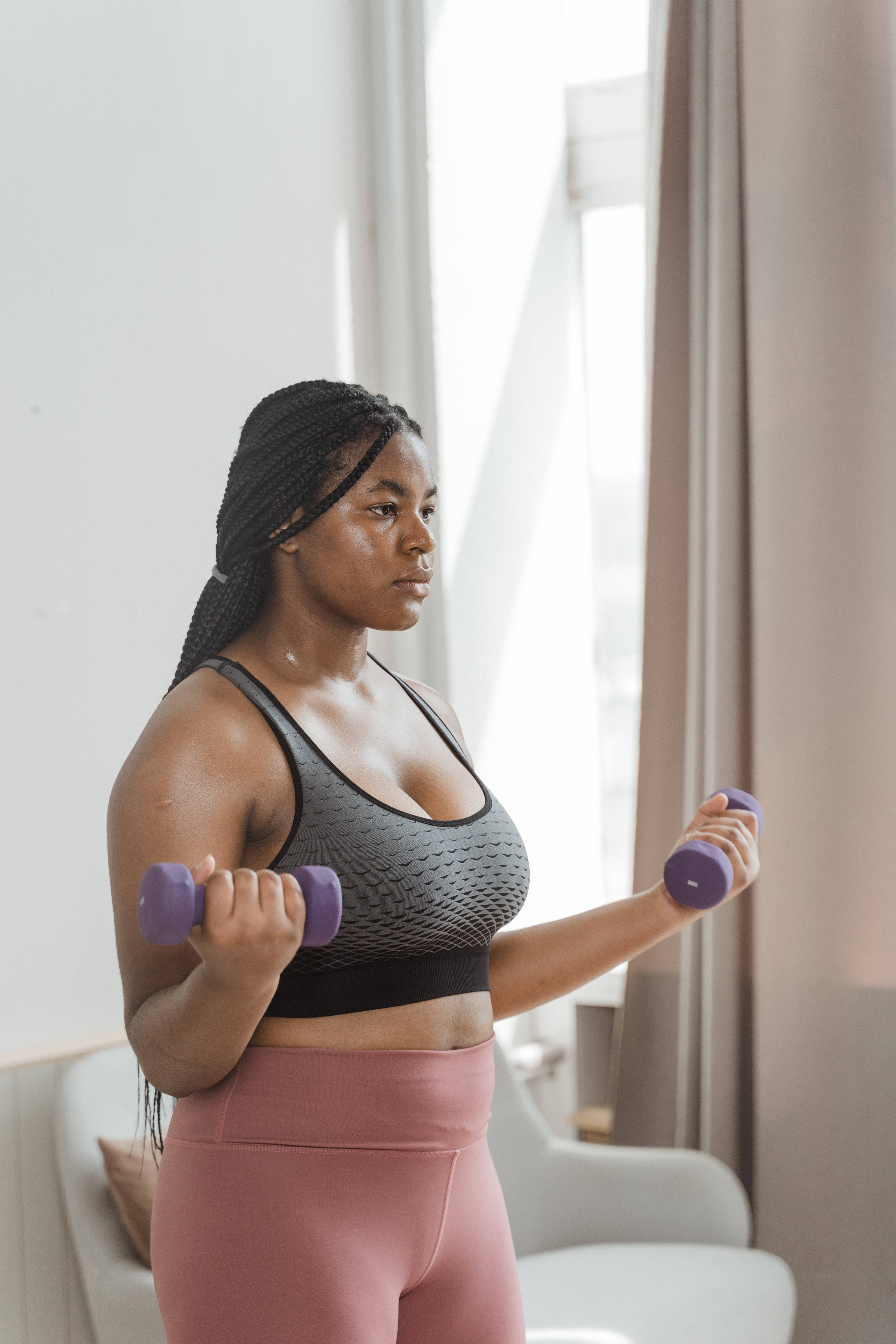 Exercise 桌布 壁紙 — 一位專注的辮子女性在明亮陽光房間內舉起紫色啞鈴，房間有米色窗簾。