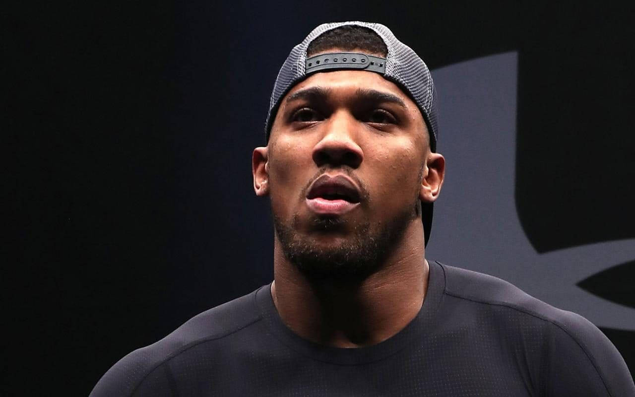 Anthony Joshua 桌布 壁紙 — 一位專注的運動員戴著灰色帽子和黑色襯衫，在帶有標誌的深色背景下凝視著。