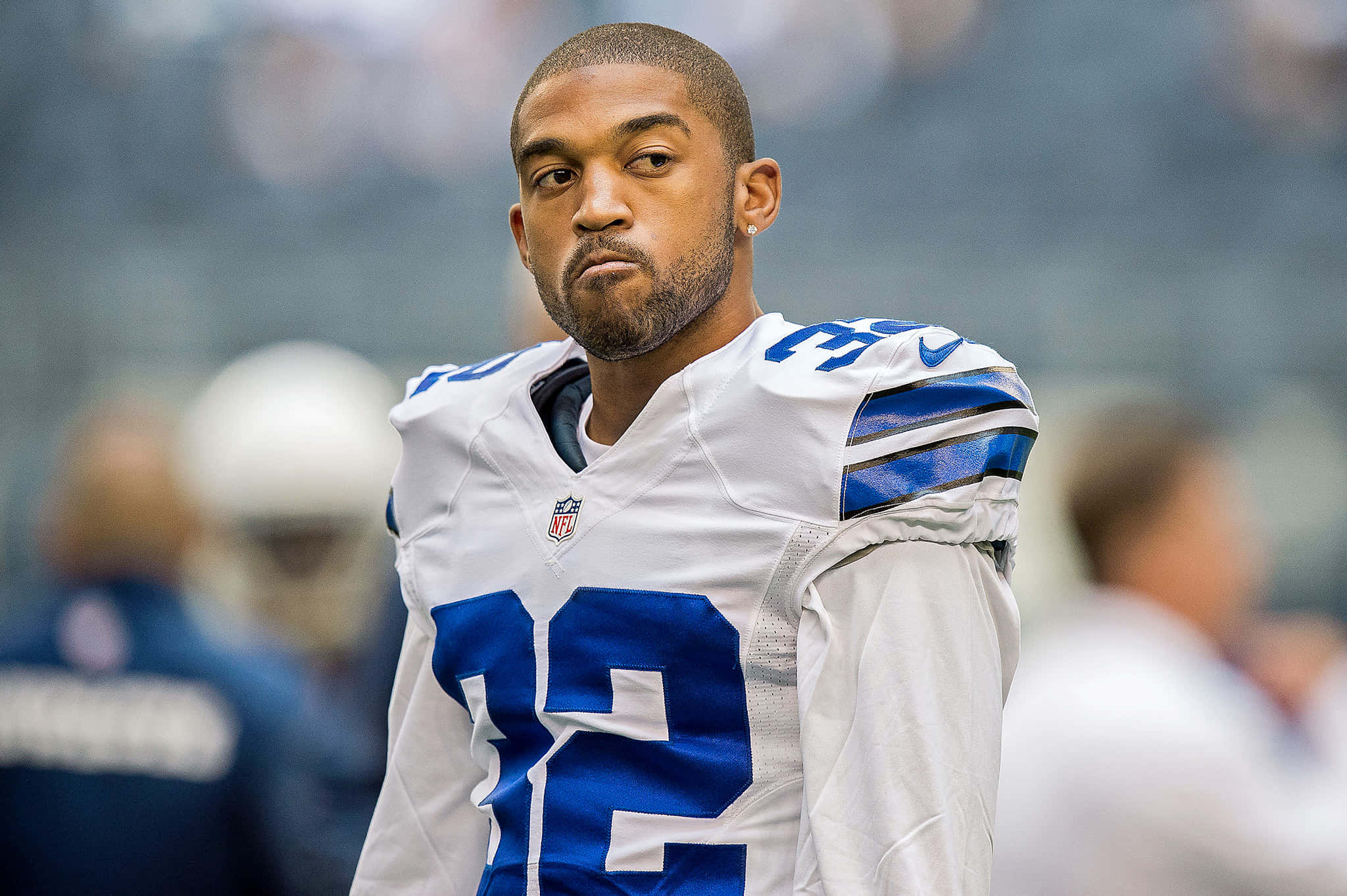 Dallas Cowboys Players 桌布 壁紙 — 一位專注的達拉斯牛仔隊美式足球員，身穿白色和藍色球衣，站在球場上，背景的體育館模糊不清。
