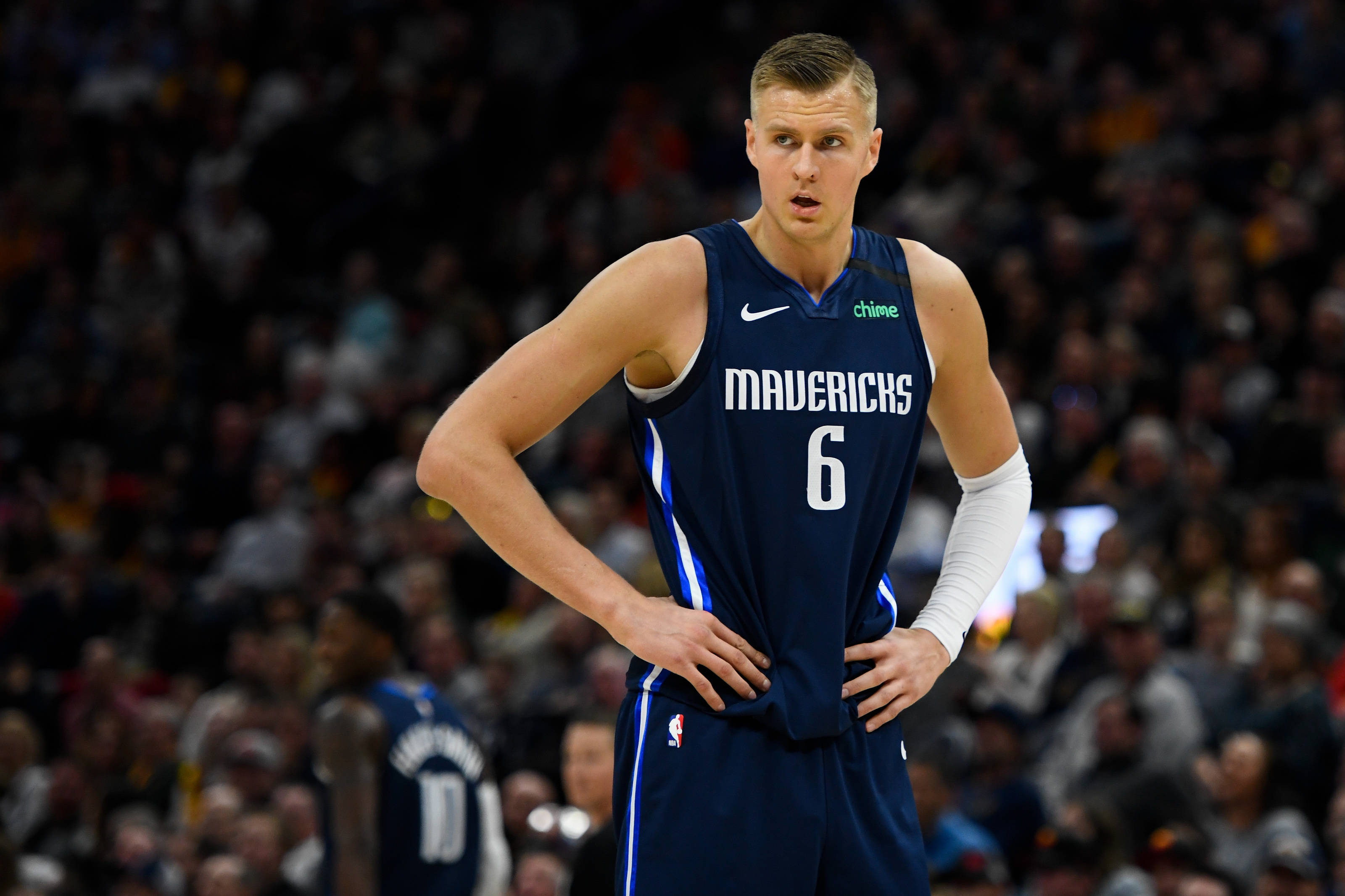Kristaps Porzingis 桌布 壁紙 — 一位專注的達拉斯獨行俠籃球運動員，身穿深藍色球衣，背號6，在明亮的場館燈光下比賽期間雙手叉腰站立。