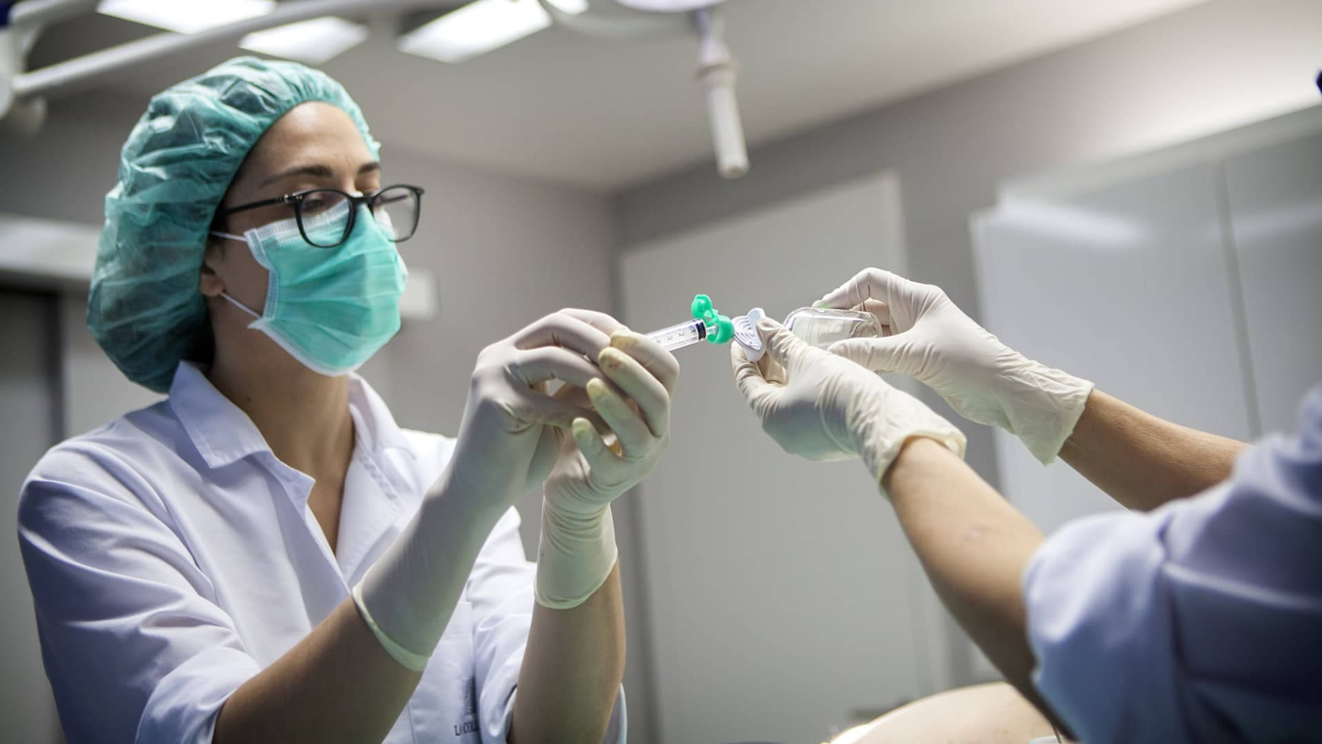Medical 桌布 壁紙 — 一位專注的醫護專業人員穿著手術衣和口罩，在明亮的臨床燈光下小心地將藥物注入注射器。