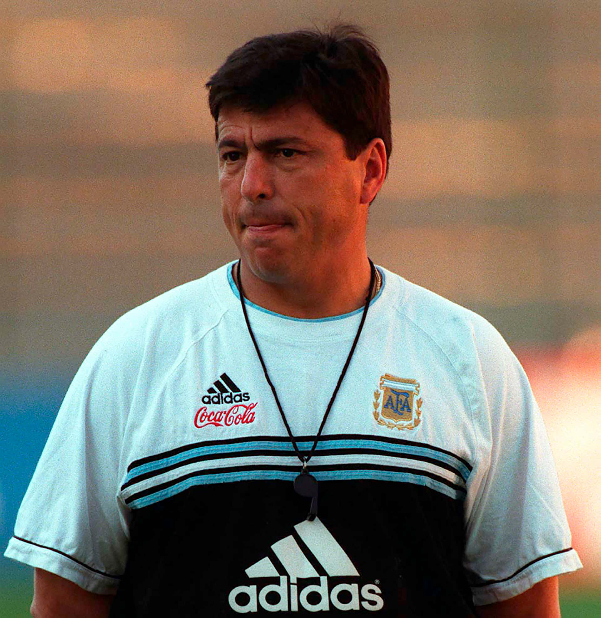 Argentina National Football Team 桌布 壁紙 — 一位專注的阿根廷足球教練，身穿淺藍色和黑色的Adidas訓練球衣，帶有可口可樂標誌，脖子上掛著哨子。