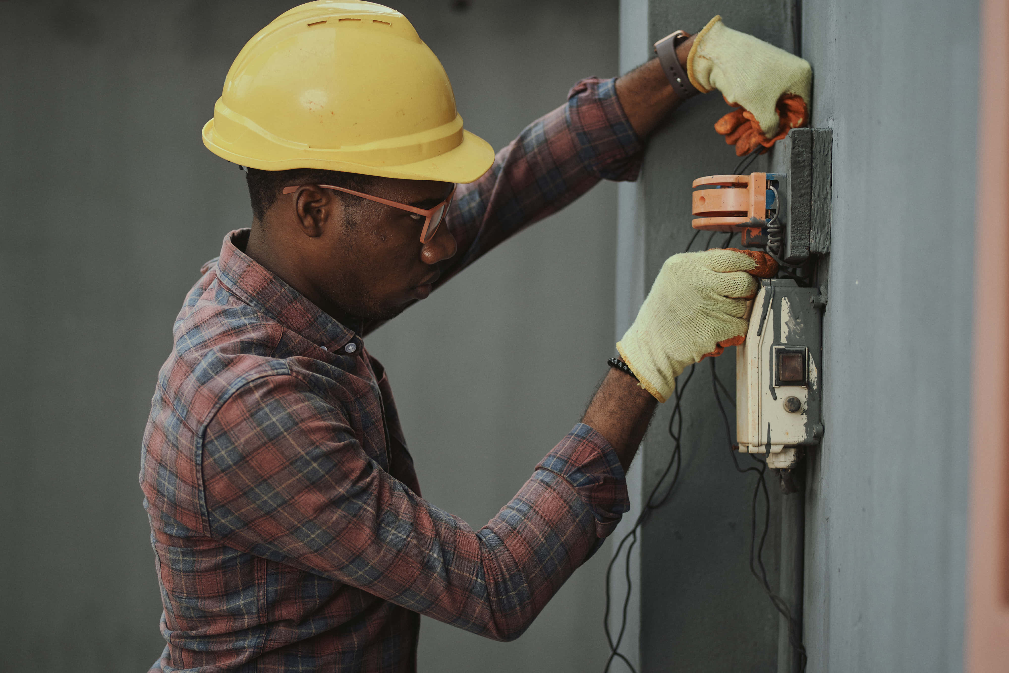 Plumbing 桌布 壁紙 — 一位專注的電工，戴著黃色頭盔和手套，正在小心地處理安裝在灰色牆上的電箱。