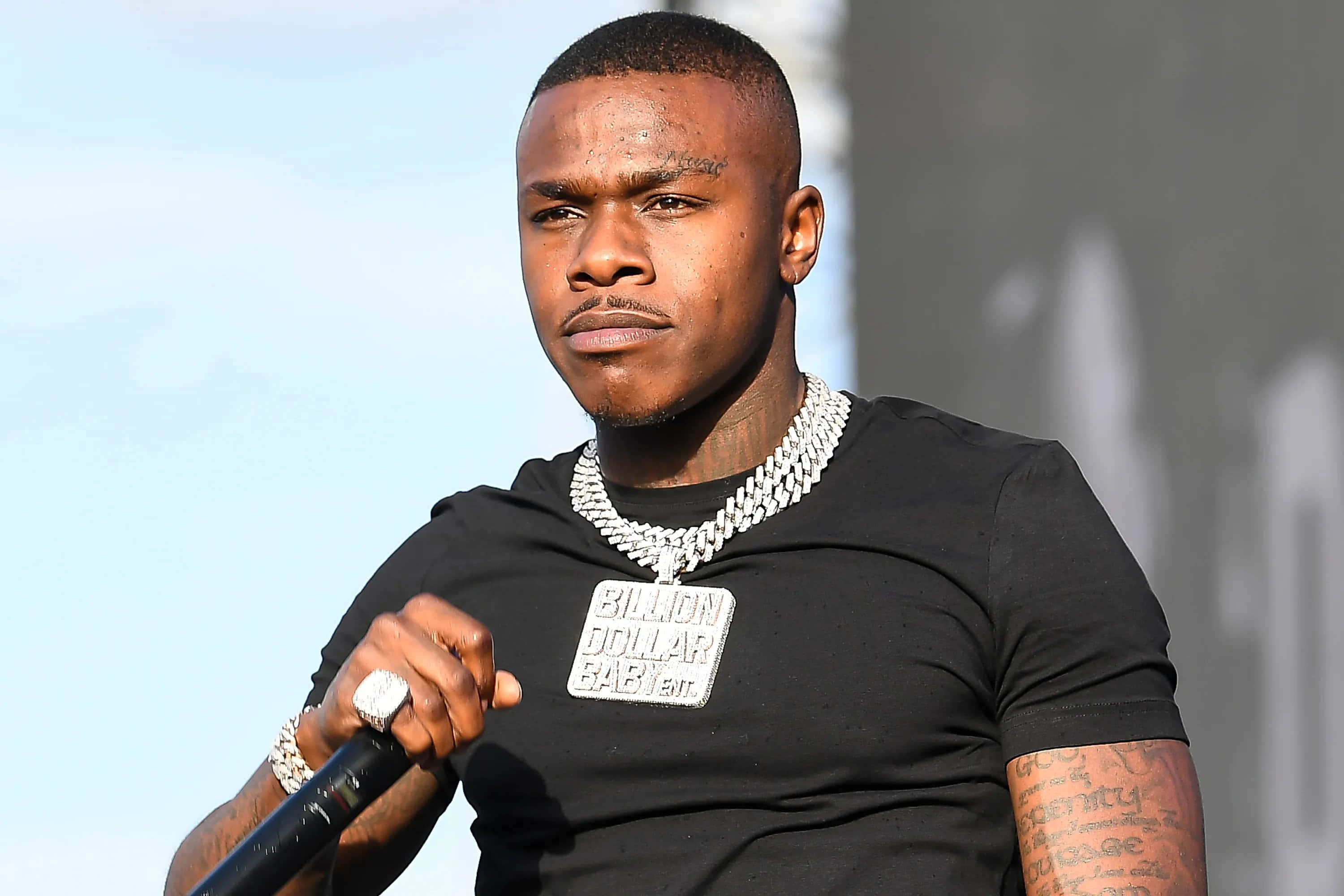 Dababy 桌布 壁紙 — 一位專注的饒舌歌手，身上有刺青和一條粗鏈，在明亮的日光下拿著麥克風在舞台上表演。