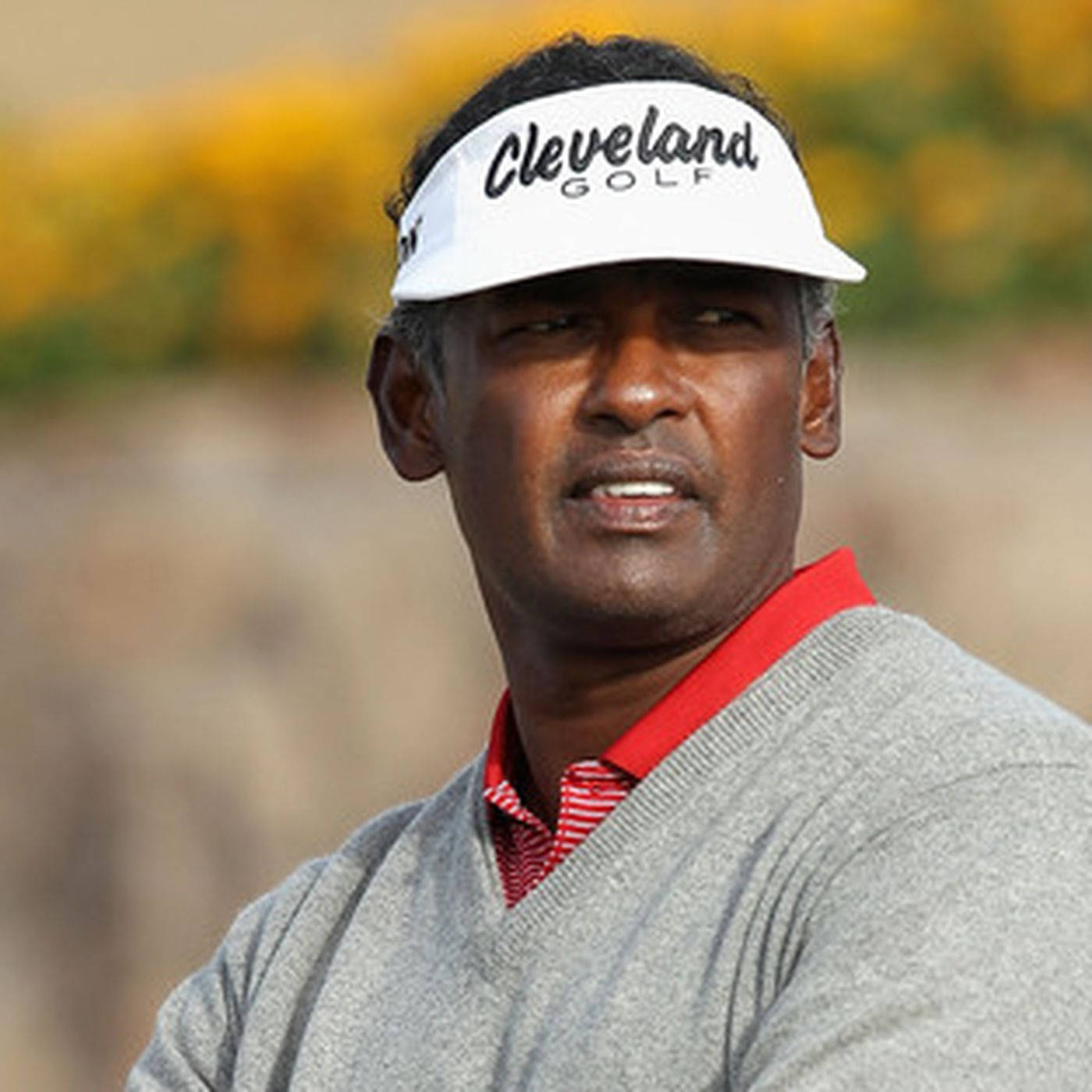 高爾夫 桌布 壁紙 — 一位專注的高爾夫球手，戴著白色 Cleveland Golf 遮陽帽和灰色毛衣，背景是模糊的黃色花卉。