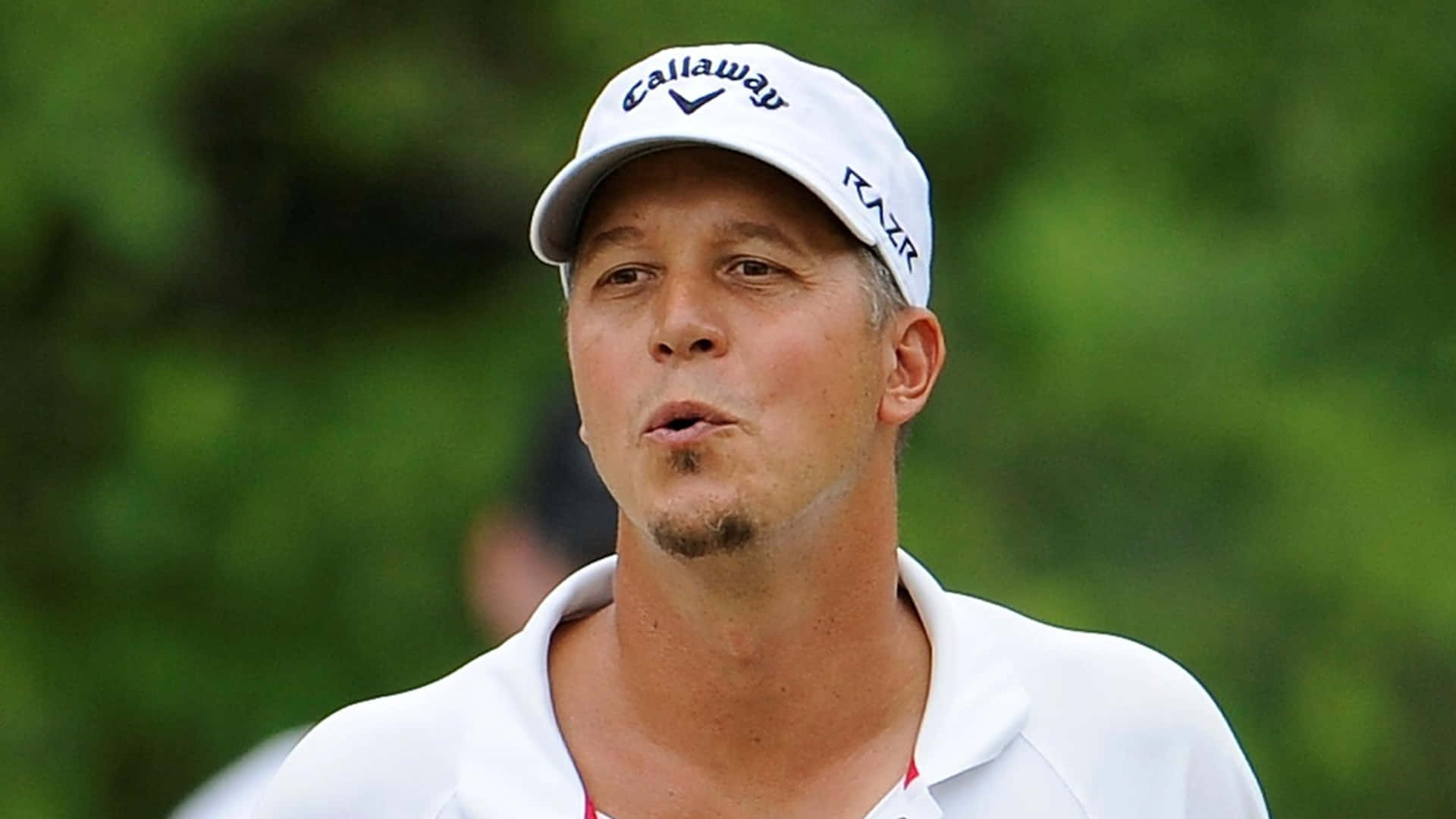  壁紙 — 一位專注的高爾夫球手，戴著白色 Callaway 帽子和 Polo 衫，站在模糊的綠色背景前。