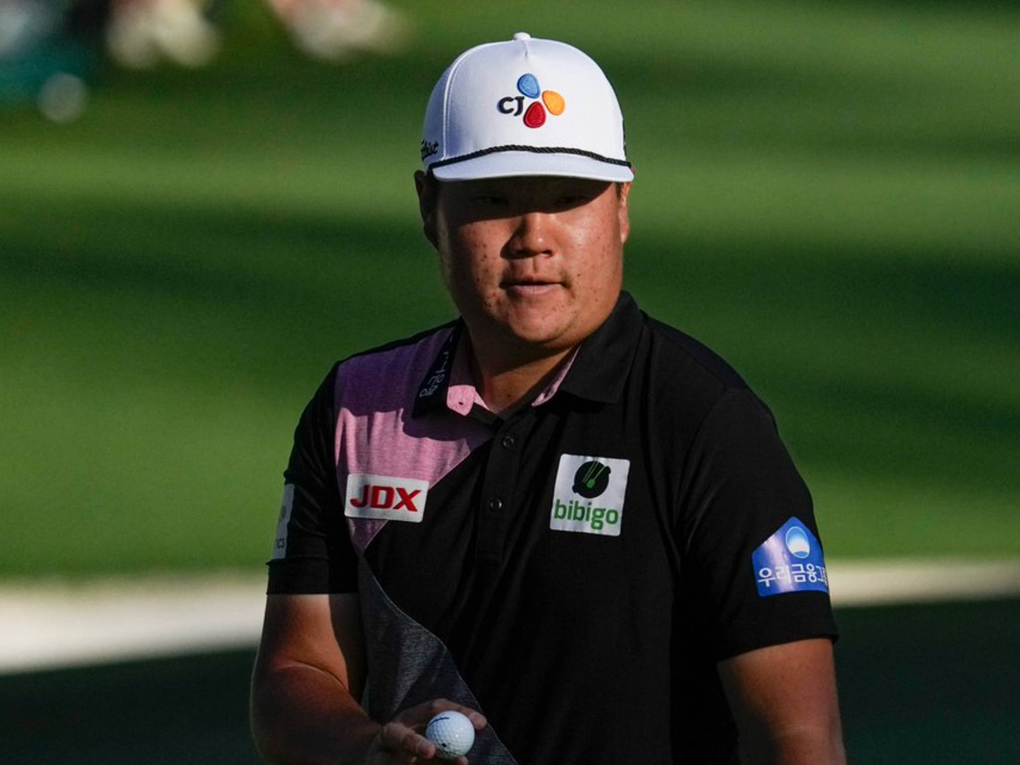 運動 桌布 壁紙 — 一位專注的高爾夫球手，戴著白色帽子，穿著黑色Polo衫，在模糊的綠色球場背景下拿著高爾夫球。