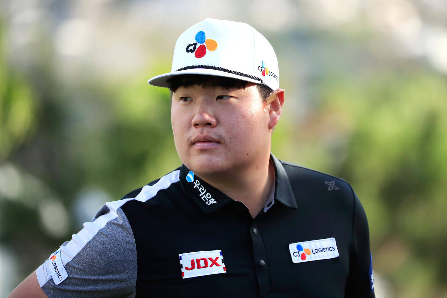運動 桌布 壁紙 — 一位專注的高爾夫球手，戴著白色帽子，穿著黑色Polo衫，站在模糊的綠色背景前。