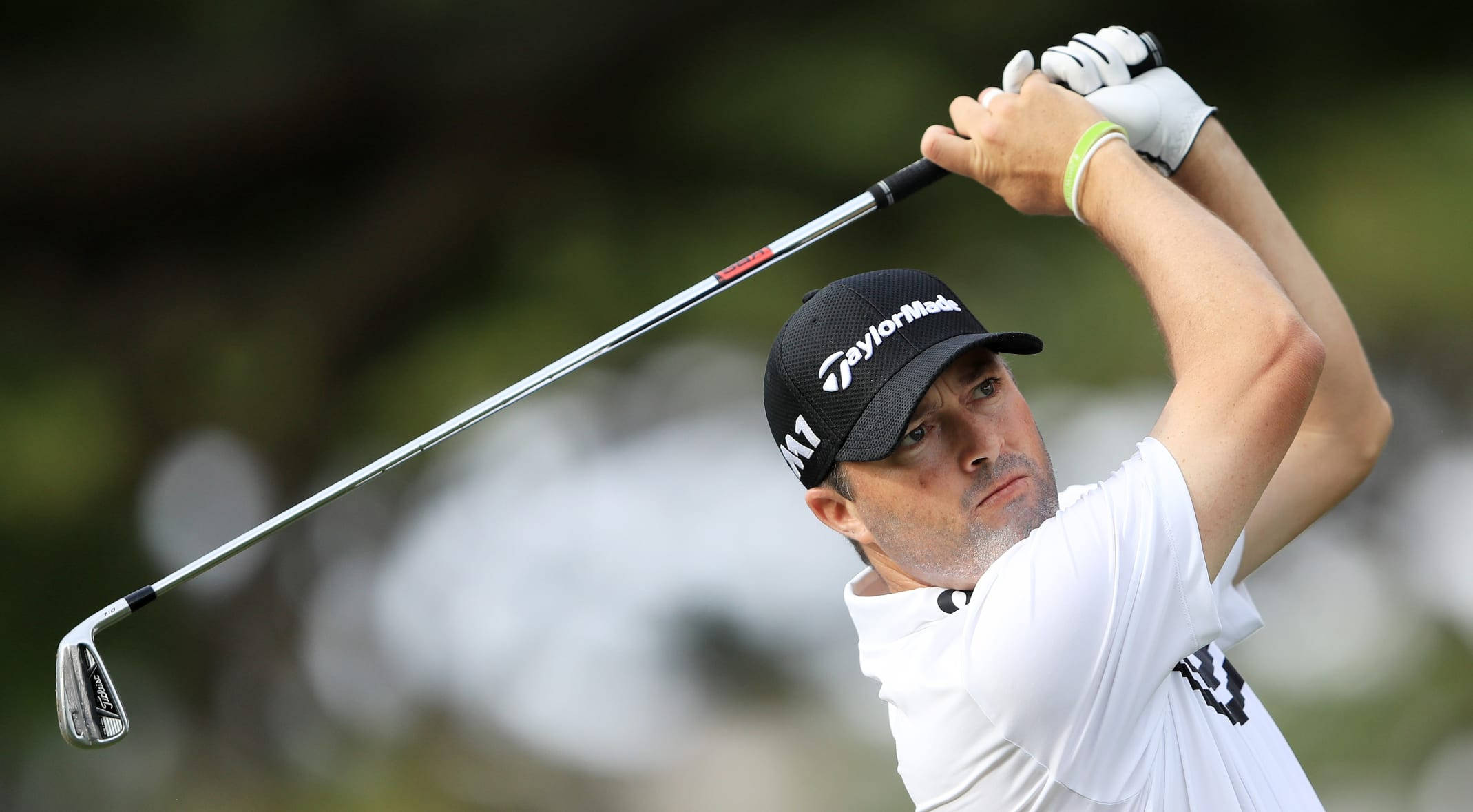 Ryan Palmer 桌布 壁紙 — 一位專注的高爾夫球手，身穿白色襯衫和黑色帽子，在模糊的綠色背景下執行強力揮桿。