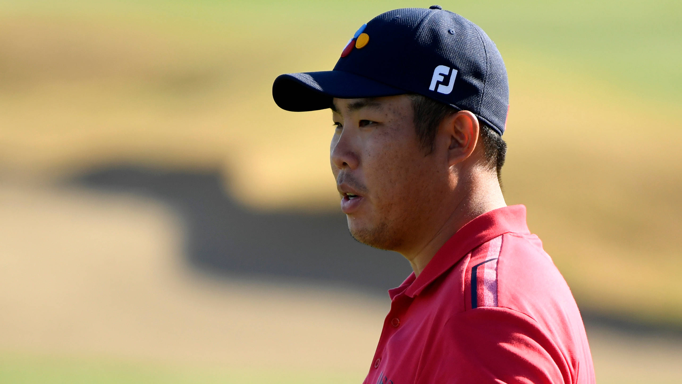 運動 桌布 壁紙 — 一位專注的高爾夫球手，身穿紅色Polo衫和深藍色帽子，站在陽光明媚的球場上，身後是模糊的果嶺和沙坑。