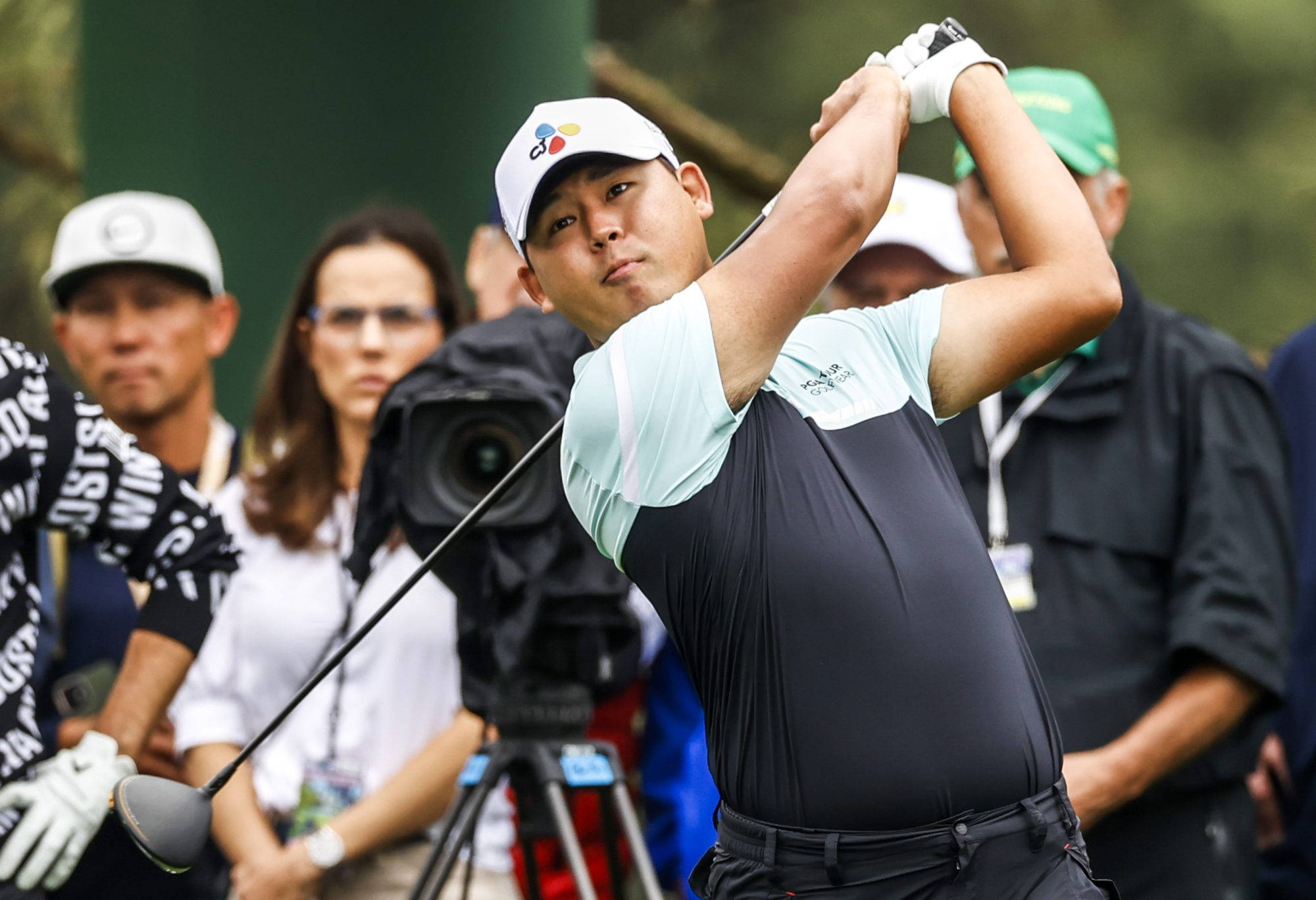 Athlete 桌布 壁紙 — 一位專注的高爾夫球手在茂密的綠色球場上揮桿，背景有觀眾和攝製組。