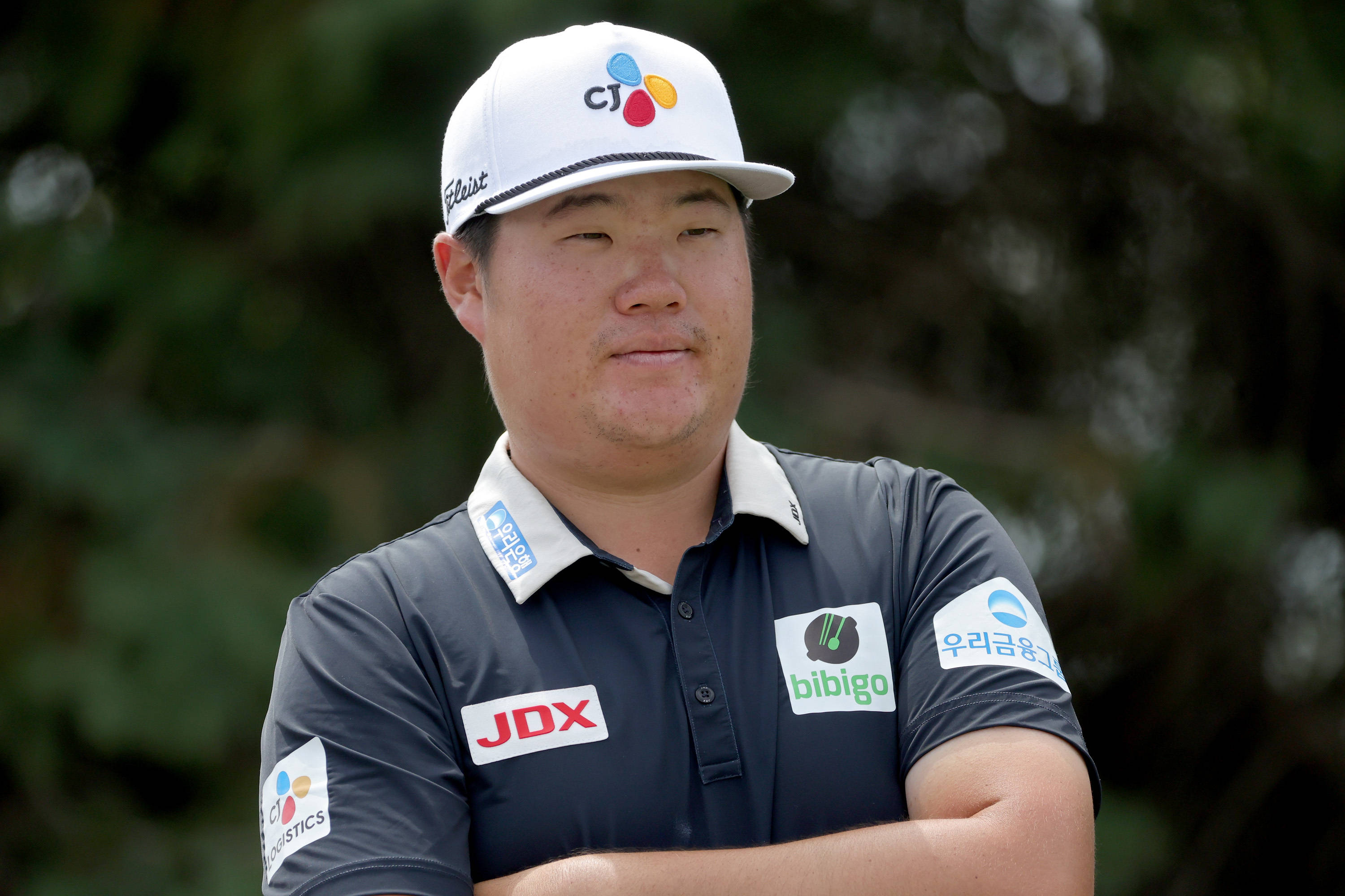 運動 桌布 壁紙 — 一位專注的高爾夫球手戴著品牌帽和Polo衫，雙臂交叉站在模糊的綠色背景前。