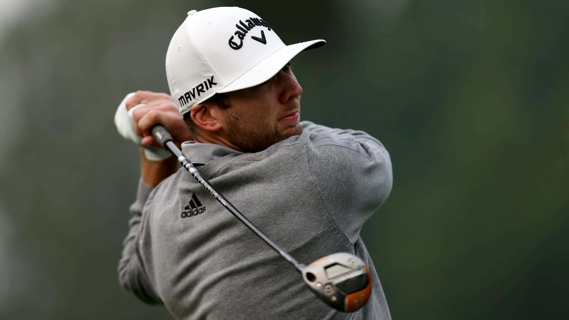  壁紙 — 一位專注的高爾夫球手正在揮桿，戴著白色 Callaway 帽子和灰色 Adidas 襯衫，背景是模糊的綠色。