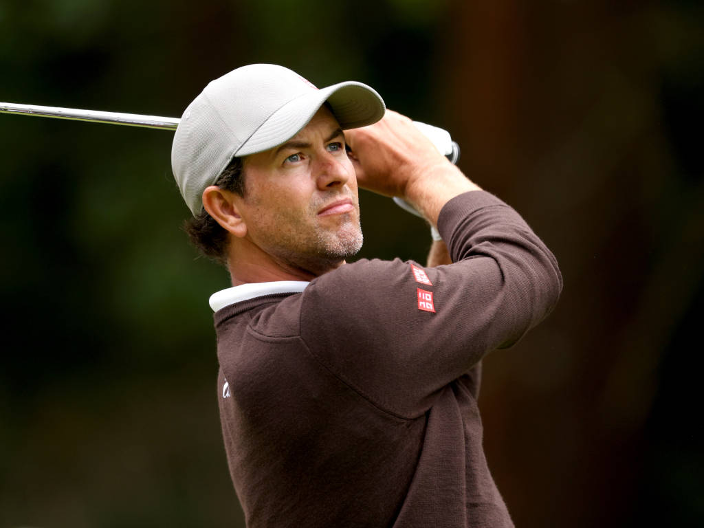 adam scott 桌布 壁紙 — 一位專注的高爾夫球手正在揮桿，戴著白色帽子，穿著棕色毛衣，背景是模糊的森林。
