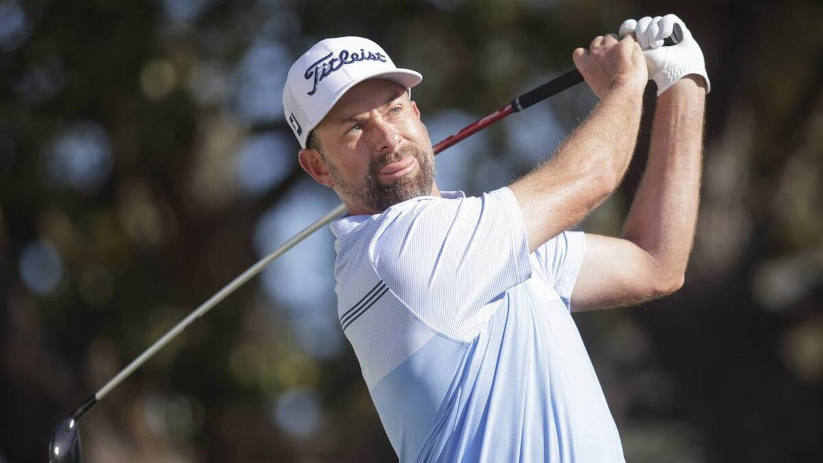  壁紙 — 一位專注的高爾夫球手正在揮桿，戴著 Titleist 帽子，身穿淺藍色襯衫，背景是模糊的樹木。