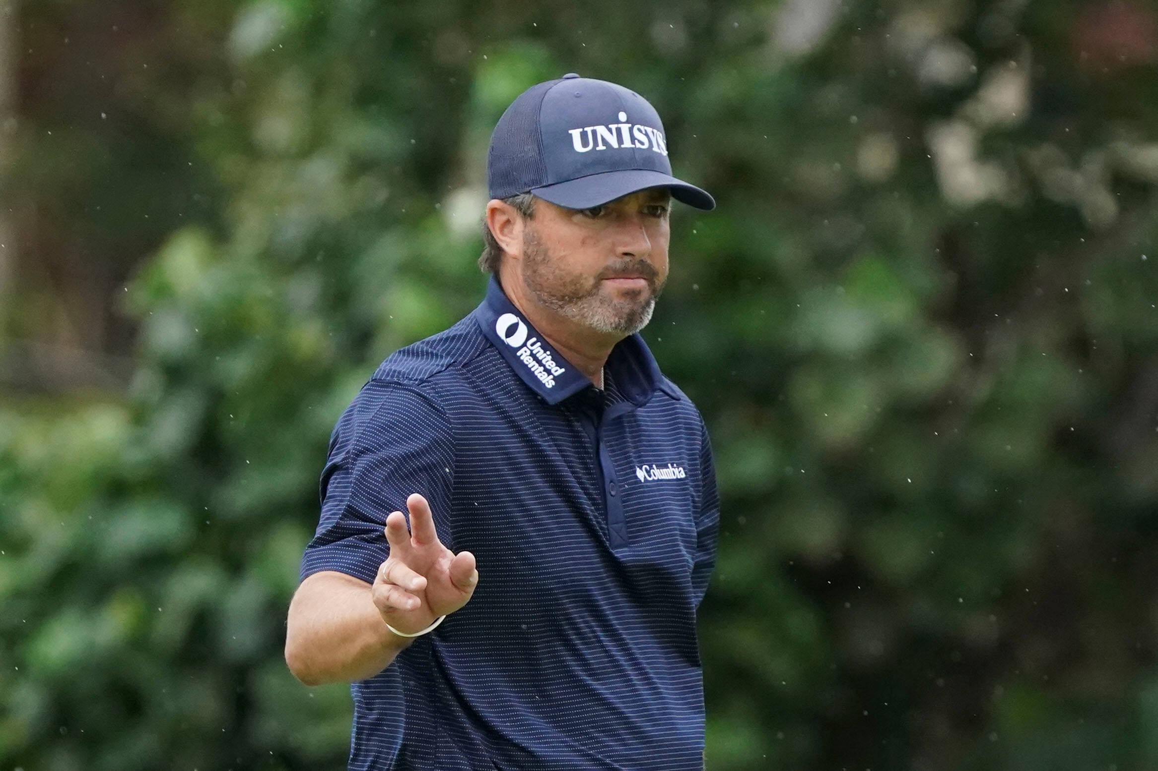 Ryan Palmer 桌布 壁紙 — 一位專注的高爾夫球手穿著海軍藍條紋Polo衫和帽子，在雨中高爾夫球場上揮桿中途做手勢，身後有模糊的綠色樹葉。