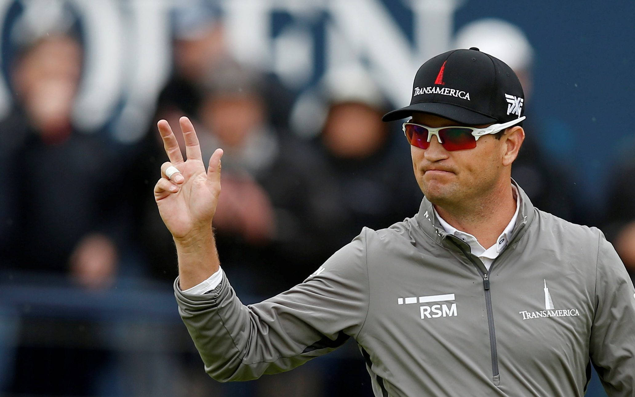 Celebrity 桌布 壁紙 — 一位專注的高爾夫球手穿著灰色外套和帽子，在陰天向人群比出和平手勢。