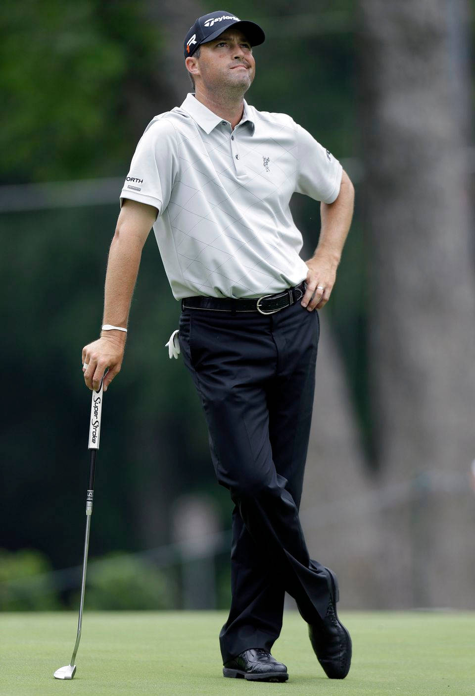 Ryan Palmer 桌布 壁紙 — 一位專注的高爾夫球手穿著白色Polo衫和黑色長褲，站在綠色高爾夫球場上，手握推桿，表情若有所思。