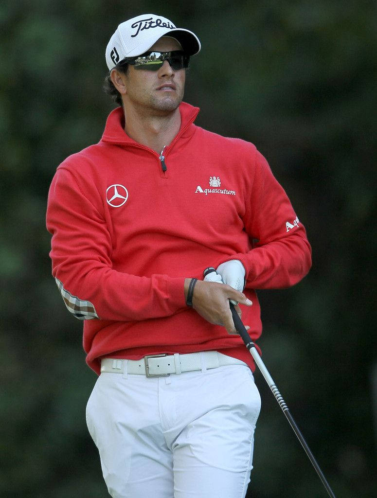adam scott 桌布 壁紙 — 一位專注的高爾夫球手穿著紅色套頭衫和白色褲子，在模糊的綠色背景下握著球桿。