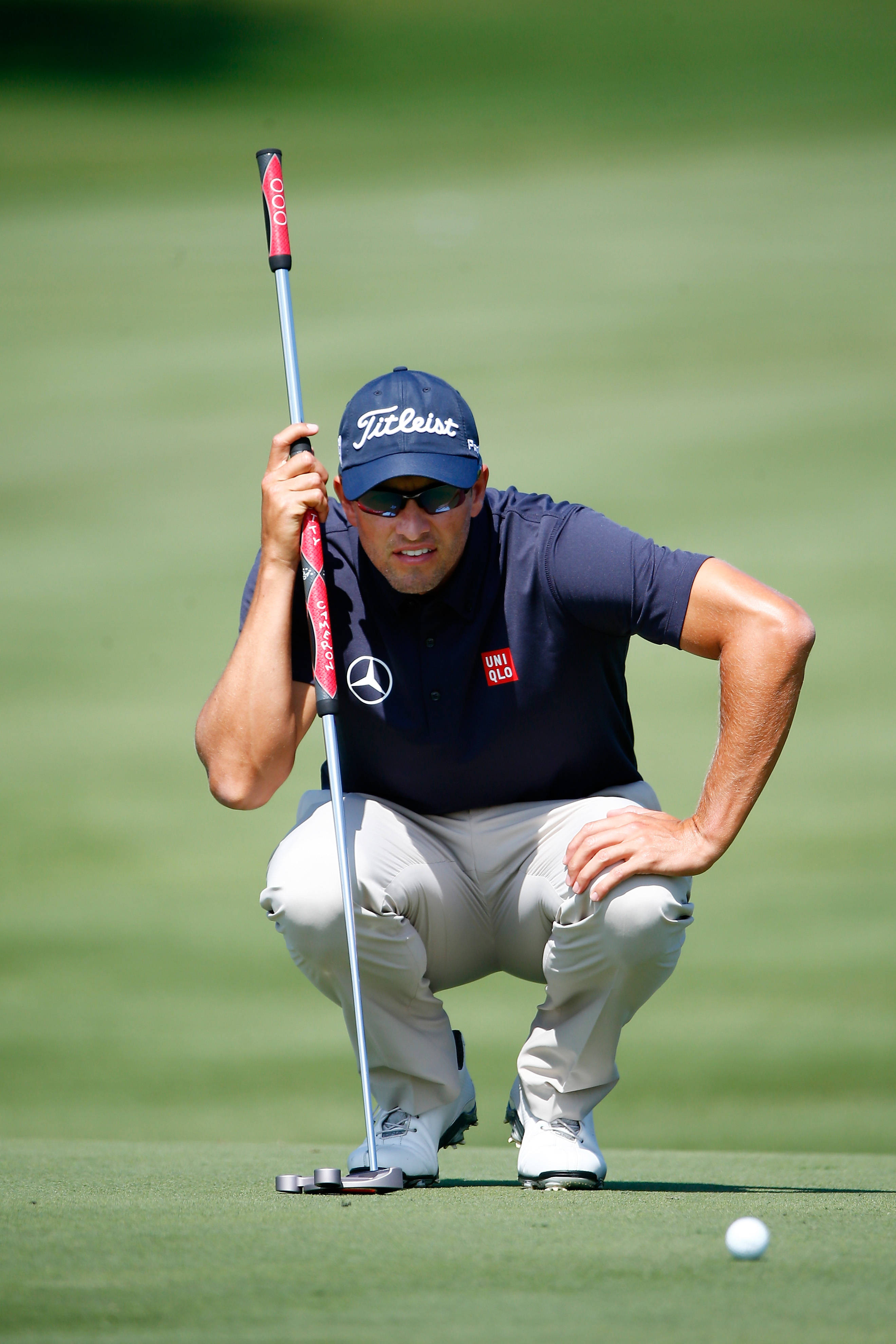 adam scott 桌布 壁紙 — 一位專注的高爾夫球手蹲在陽光普照的果嶺上，用紅色握把的推桿瞄準推桿路線。