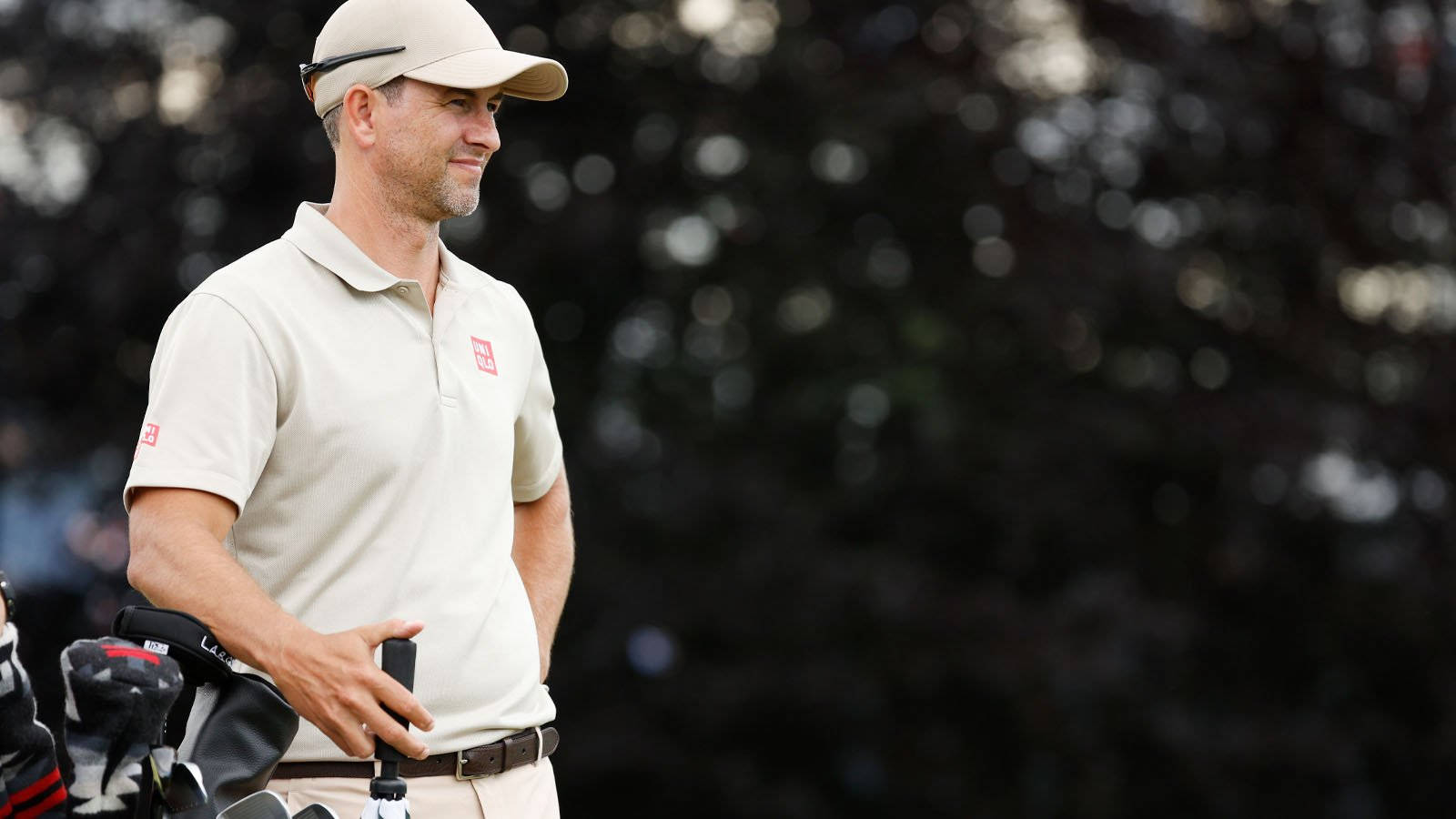 adam scott 桌布 壁紙 — 一位專注的高爾夫球手身穿米色服裝，在柔和日光下站在模糊的綠色球場上，旁邊放著他的球袋。