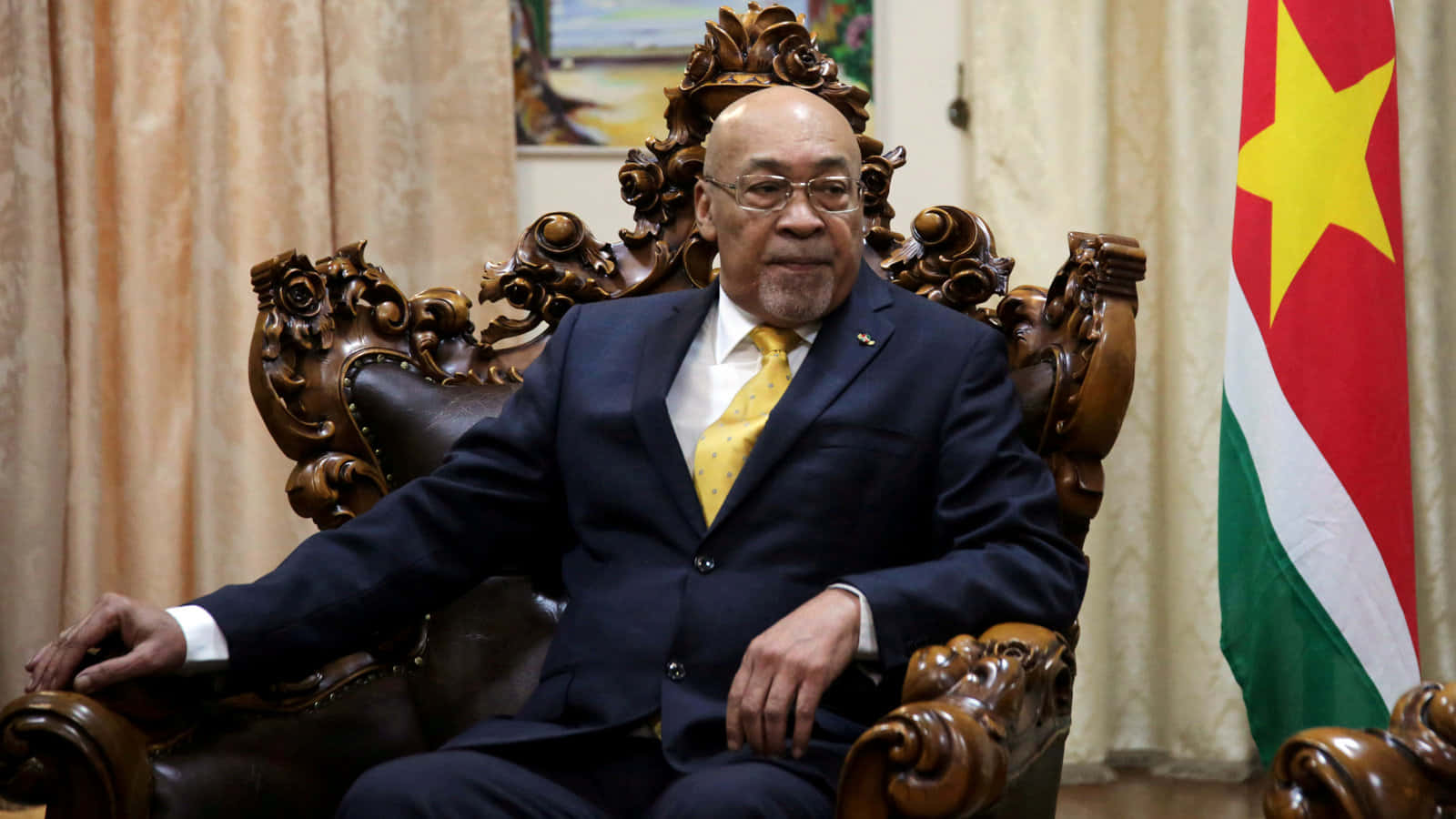 Leadership 桌布 壁紙 — 一位尊貴的男子身穿深色西裝和黃色領帶，莊嚴地坐在華麗的木製寶座椅上，旁邊有一面蘇利南國旗。