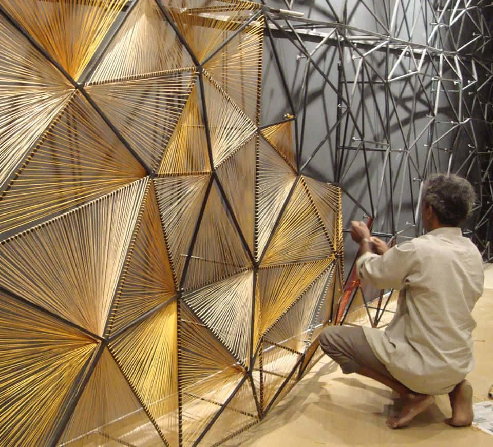 Installation Art 桌布 壁紙 — 一位工匠跪著組裝一件由金色竹條和黑色金屬框架製成的大型幾何牆壁藝術品。