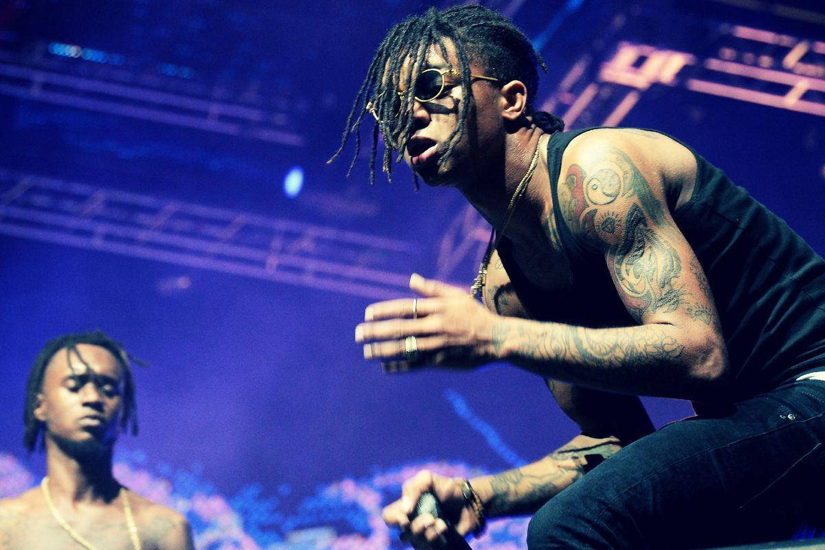 Swae Lee 桌布 壁紙 — 一位帶有刺青、梳著辮子頭和戴太陽眼鏡的饒舌歌手，在藍色燈光的舞台上充滿活力地拿著麥克風表演。