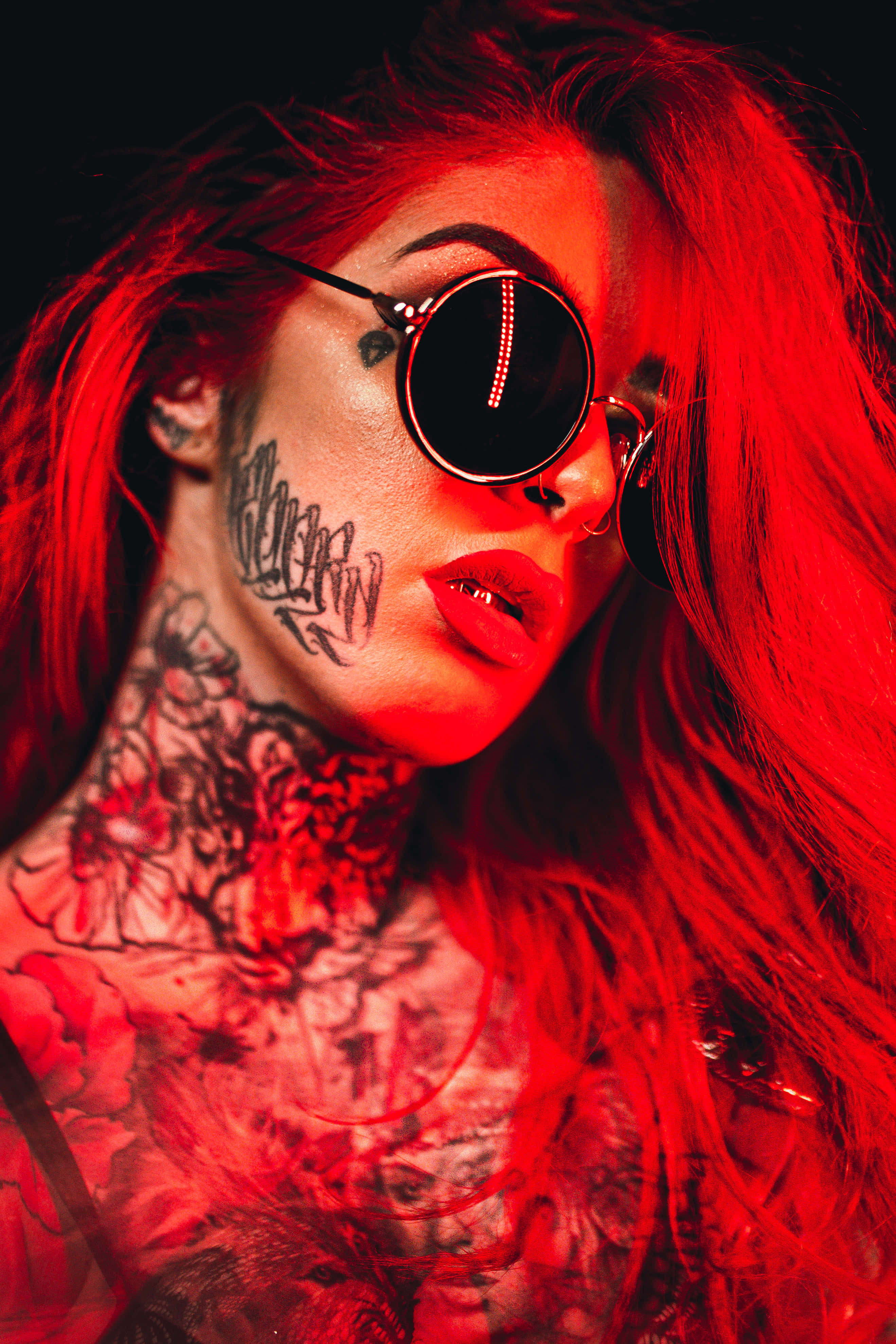 Badass Girl 桌布 壁紙 — 一位帶有刺青和圓形太陽眼鏡的女性，在黑色背景下沐浴在強烈的紅光中。
