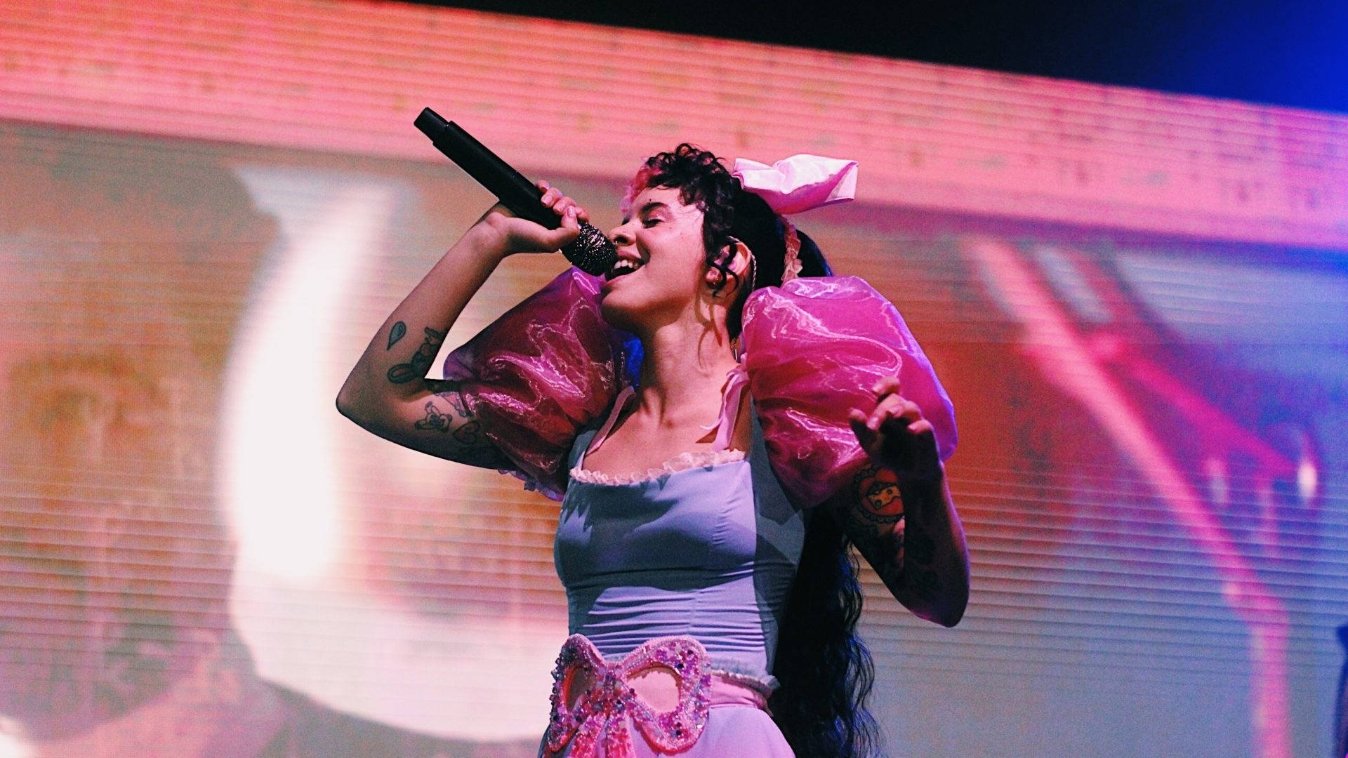 Melanie 桌布 壁紙 — 一位帶有刺青和粉紅色蝴蝶結的歌手在充滿活力的粉紅色和藍色燈光下充滿活力地在舞台上表演。