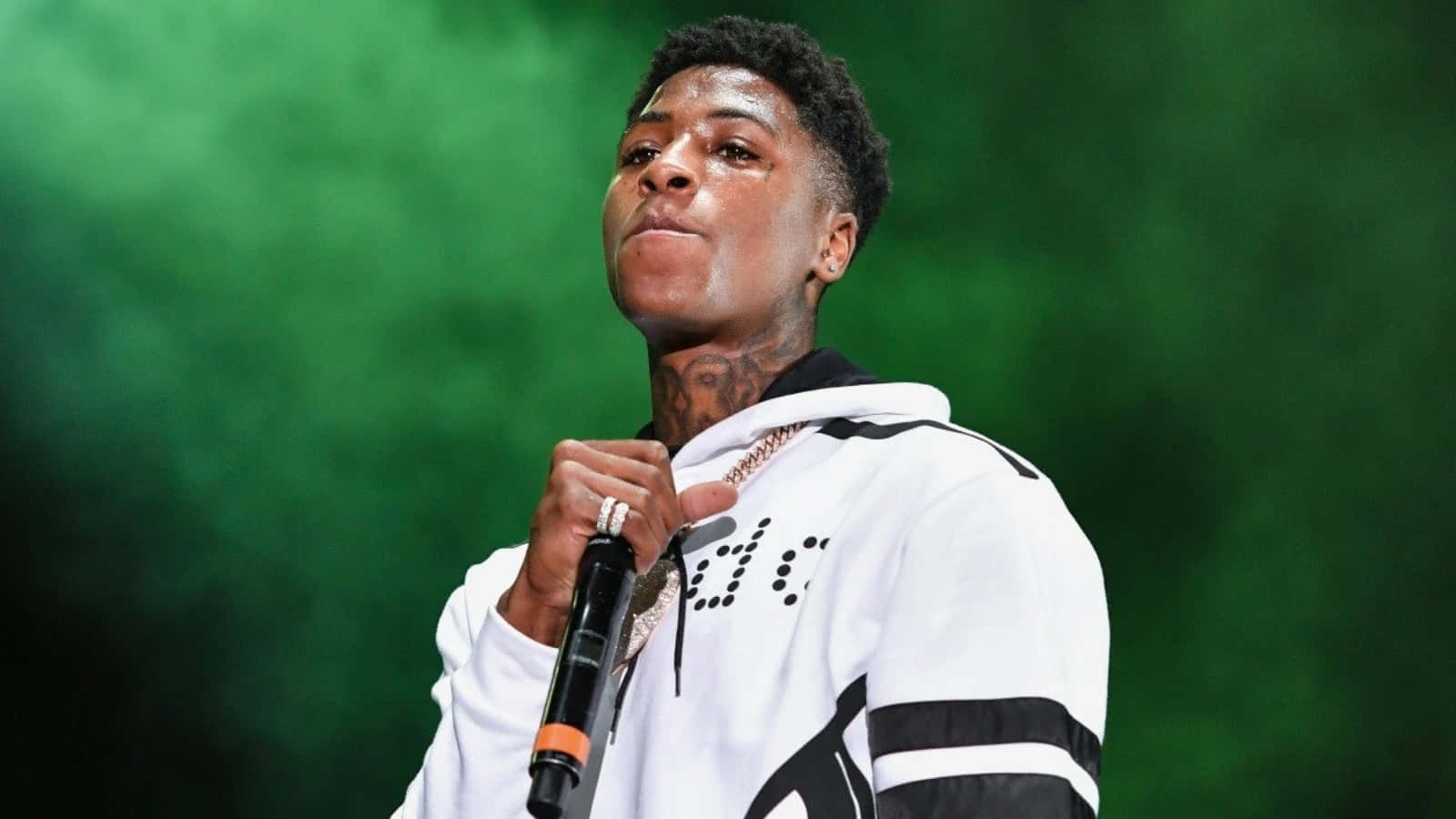 NBA YoungBoy 桌布 壁紙 — 一位帶有刺青的說唱歌手在煙霧繚繞的綠色舞台背景下手持麥克風。