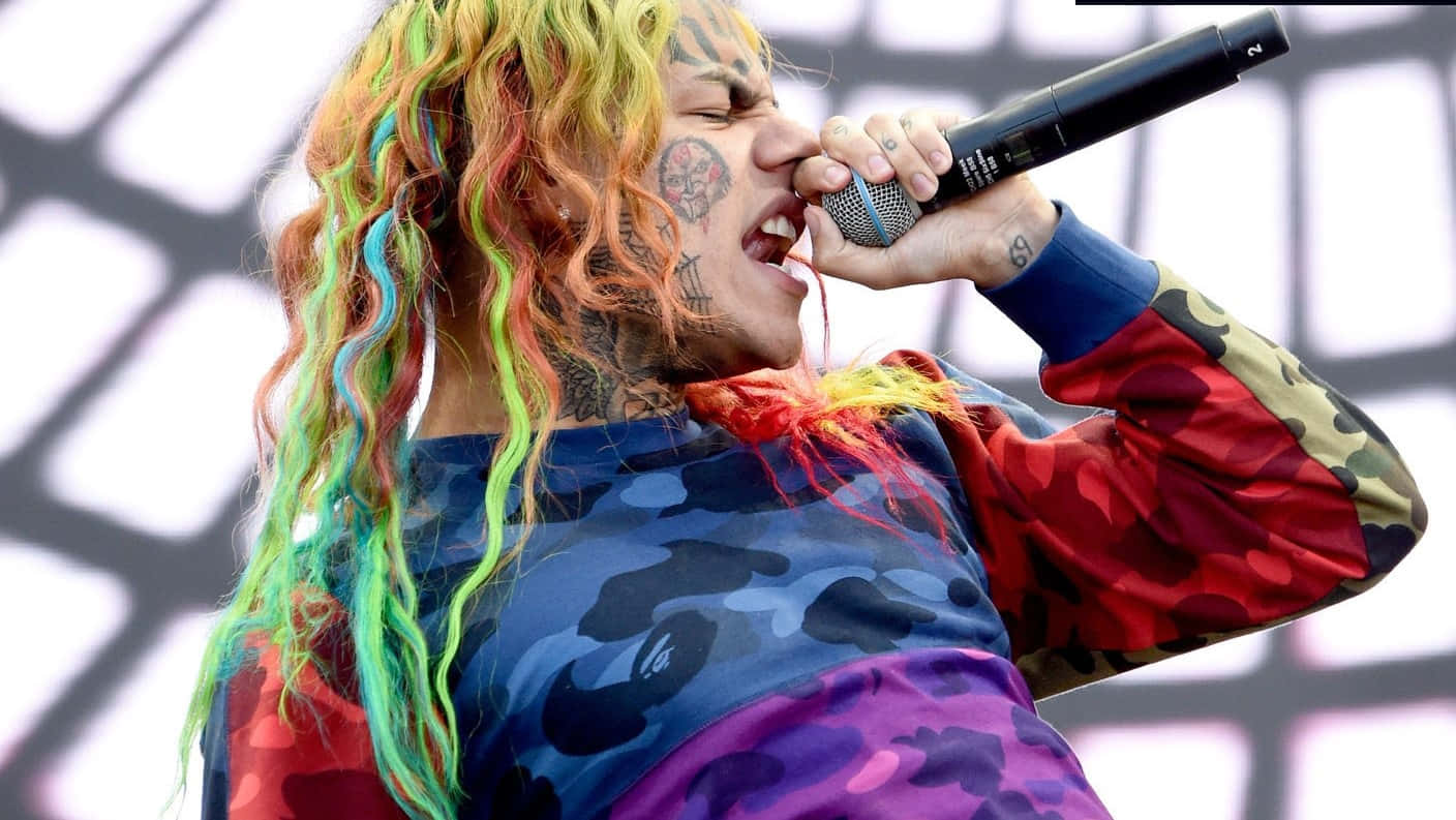 69 Rapper 桌布 壁紙 — 一位帶有彩虹色辮子和刺青的饒舌歌手，穿著色彩繽紛的迷彩外套對著麥克風大喊。