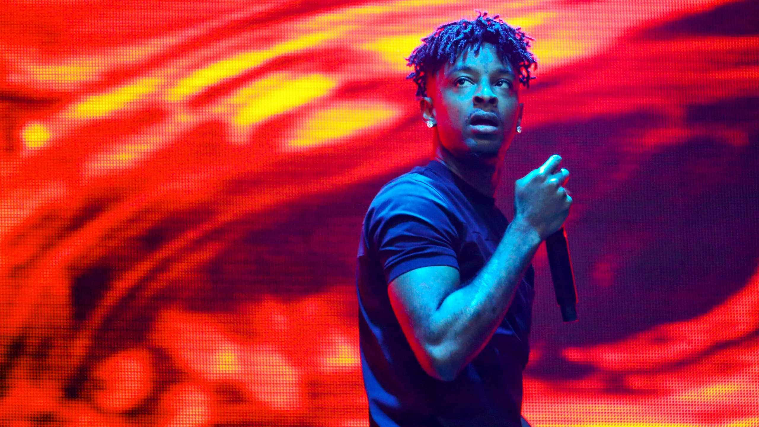 21 Savage 桌布 壁紙 — 一位帶有藍色辮子的表演者對著麥克風唱歌，背景是充滿活力的紅色和黃色數位背景。