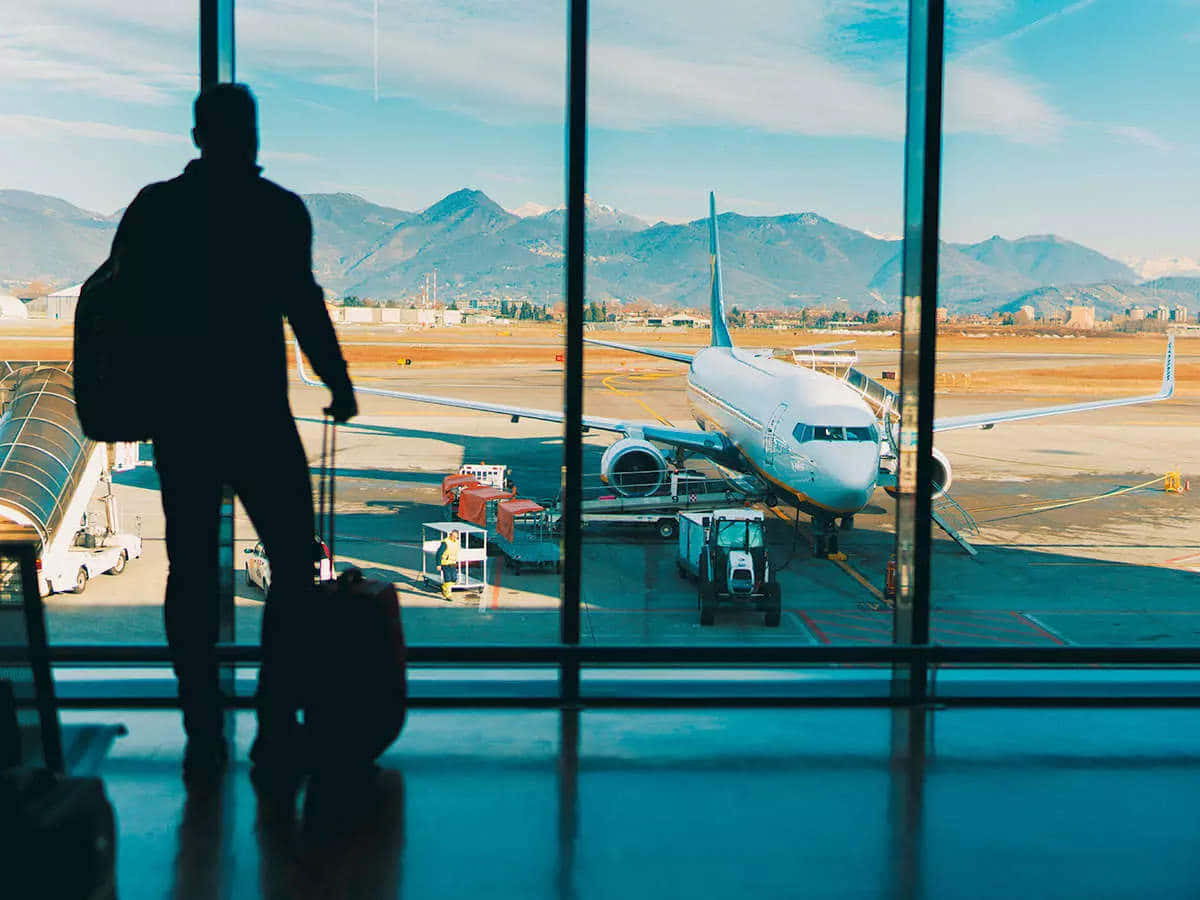Travel Desktop 桌布 壁紙 — 一位帶著行李的旅客的剪影站在機場窗前，凝視著停泊在群山背景下的飛機。