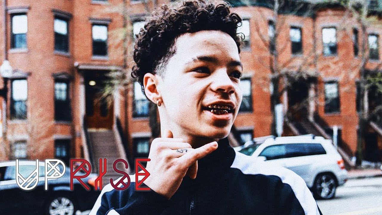 Lil Mosey 桌布 壁紙 — 一位年輕人，捲髮戴戒指，自信地指向鏡頭，背景是磚砌排屋和停泊的汽車。