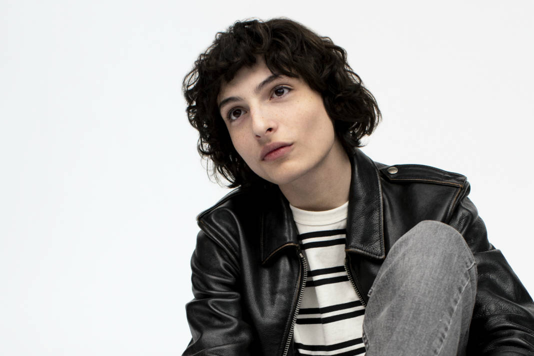 finn wolfhard 桌布 壁紙 — 一位年輕人有捲曲深色頭髮，穿著黑色皮夾克內搭條紋襯衫，背景為純白色。