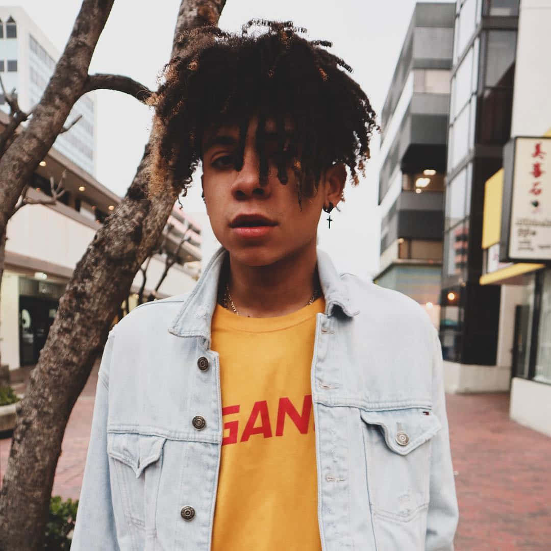Hip Hop 桌布 壁紙 — 一位年輕人有捲髮，身穿牛仔外套，站在城市人行道上，旁邊有樹，背景是現代建築。